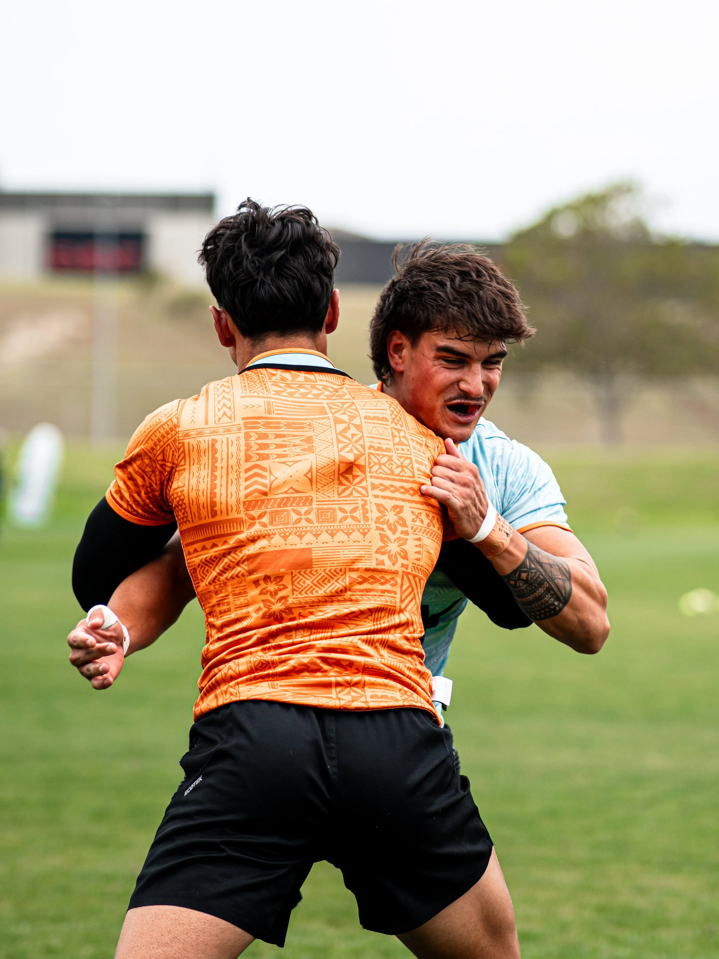 Two rugby players are training each other on a grassy field, one wearing an orange patterned jersey with black shorts and the other in a light blue jersey.