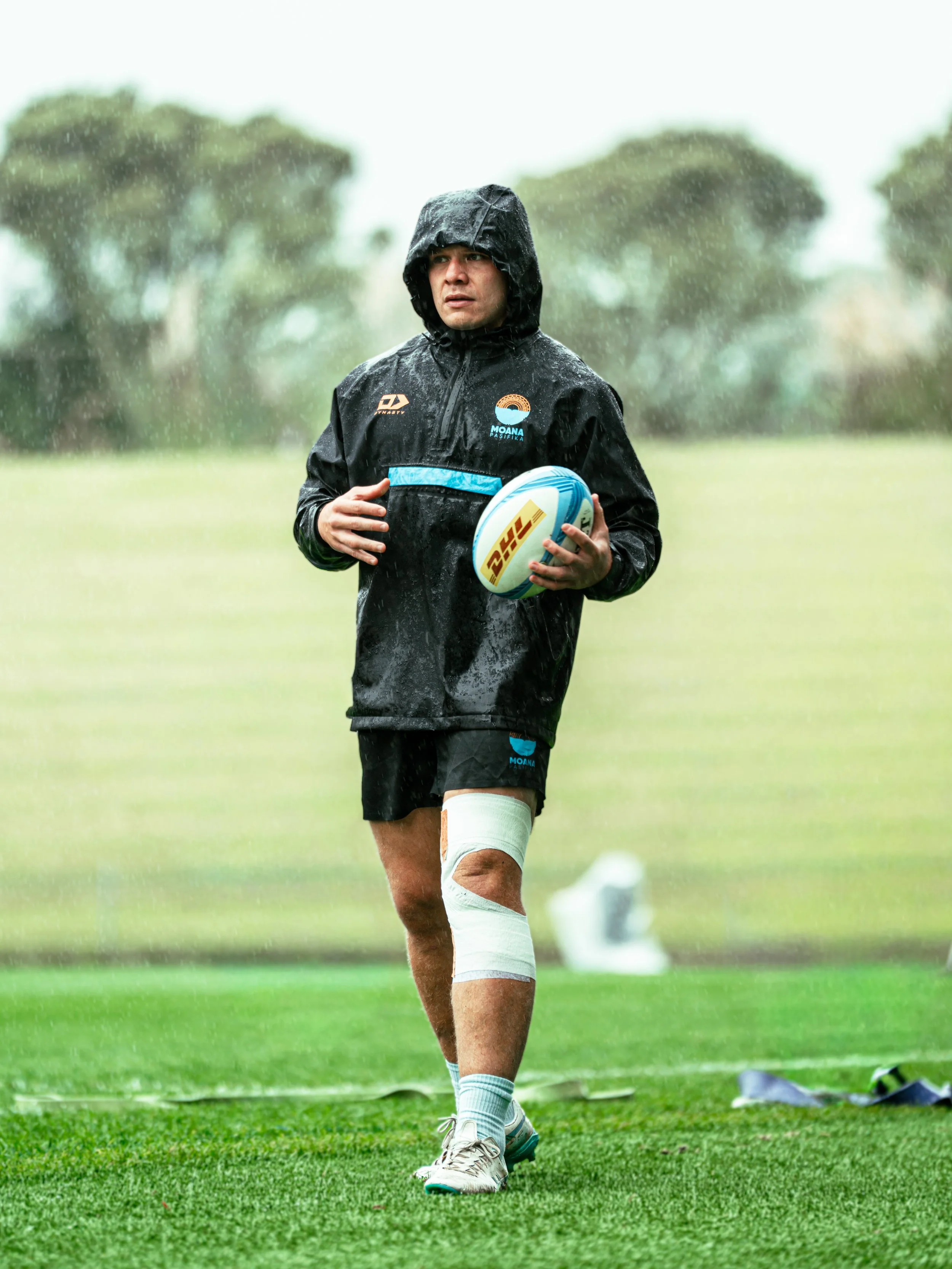 A rugby player standing on a field in pouring rain, holding a rugby ball, wearing black and blue sportswear, with a focused expression on his face.