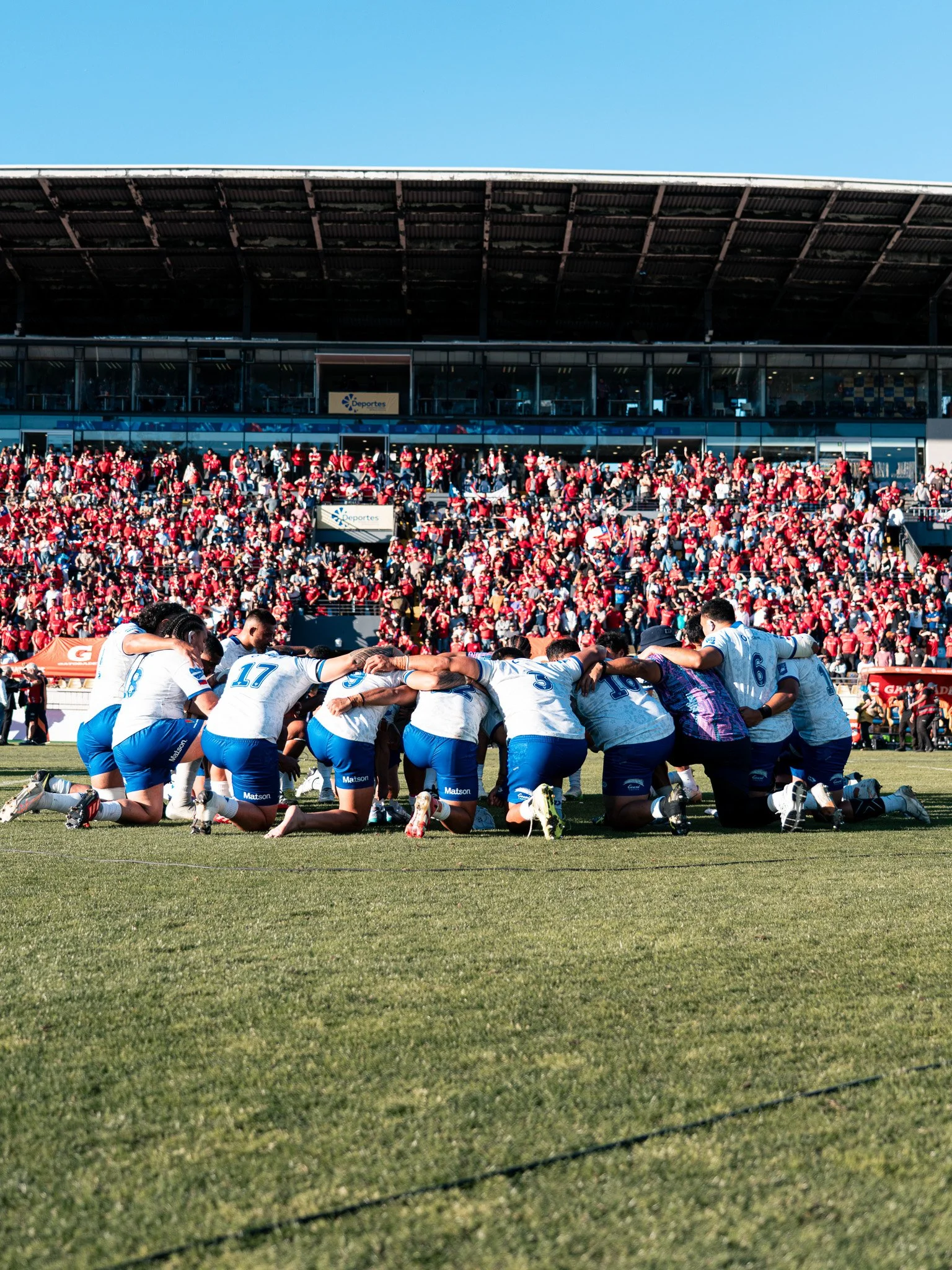 A group of rugby players kneeling and praying on the field during a match, with a large crowd of spectators in red in the stands behind them.