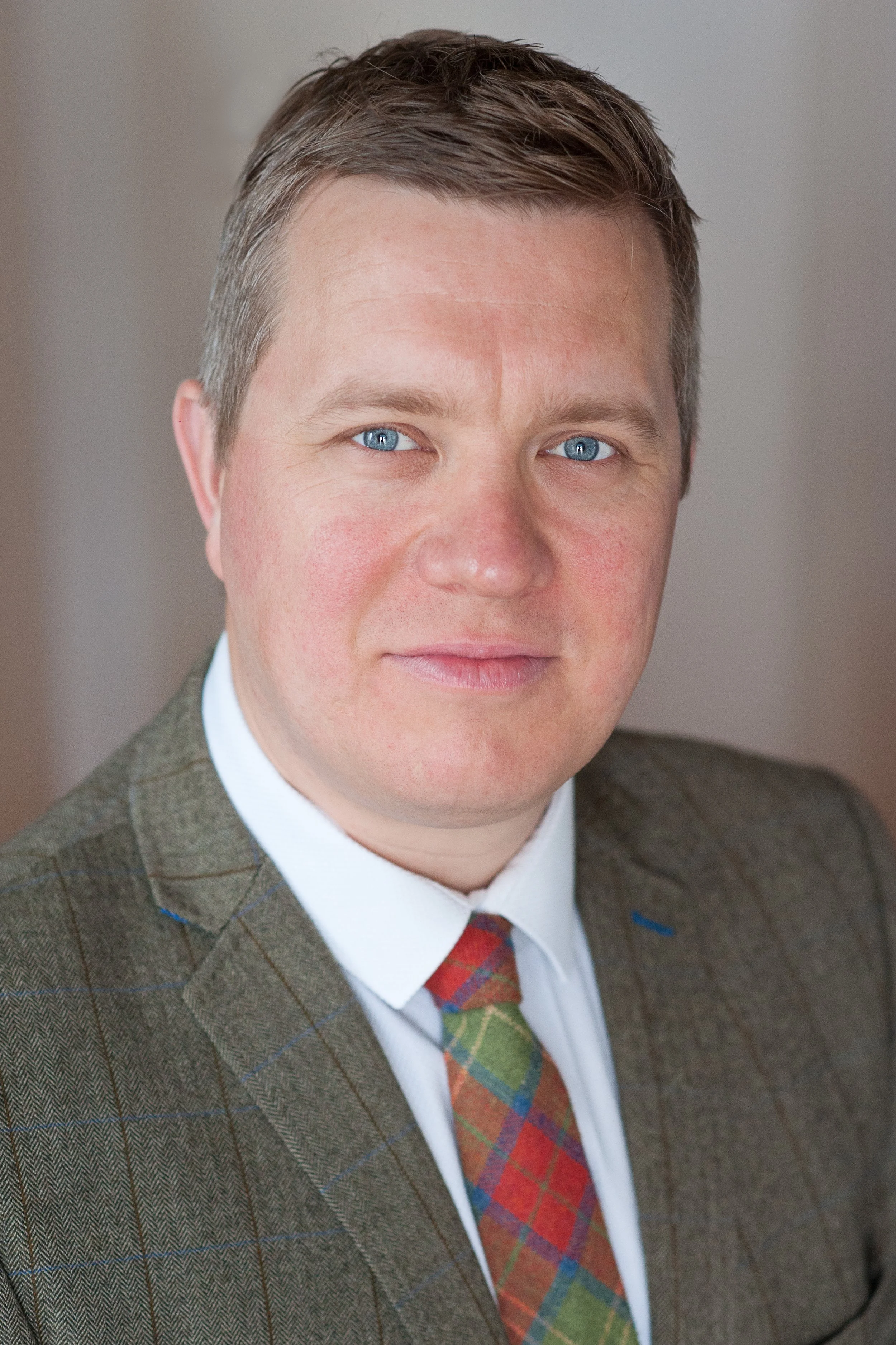 A portrait of a man with light brown hair, blue eyes, and a slight smile, wearing a brown checkered suit jacket, a white shirt, and a colorful plaid tie.