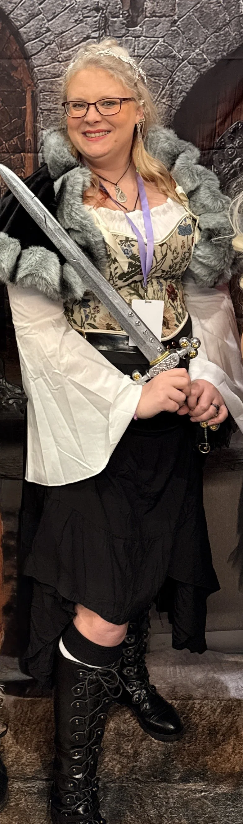 A woman dressed in medieval or fantasy costume with a gray fur cloak, floral embroidered corset, white and black skirt, and knee-high lace-up boots, holding a sword, smiling at an event.