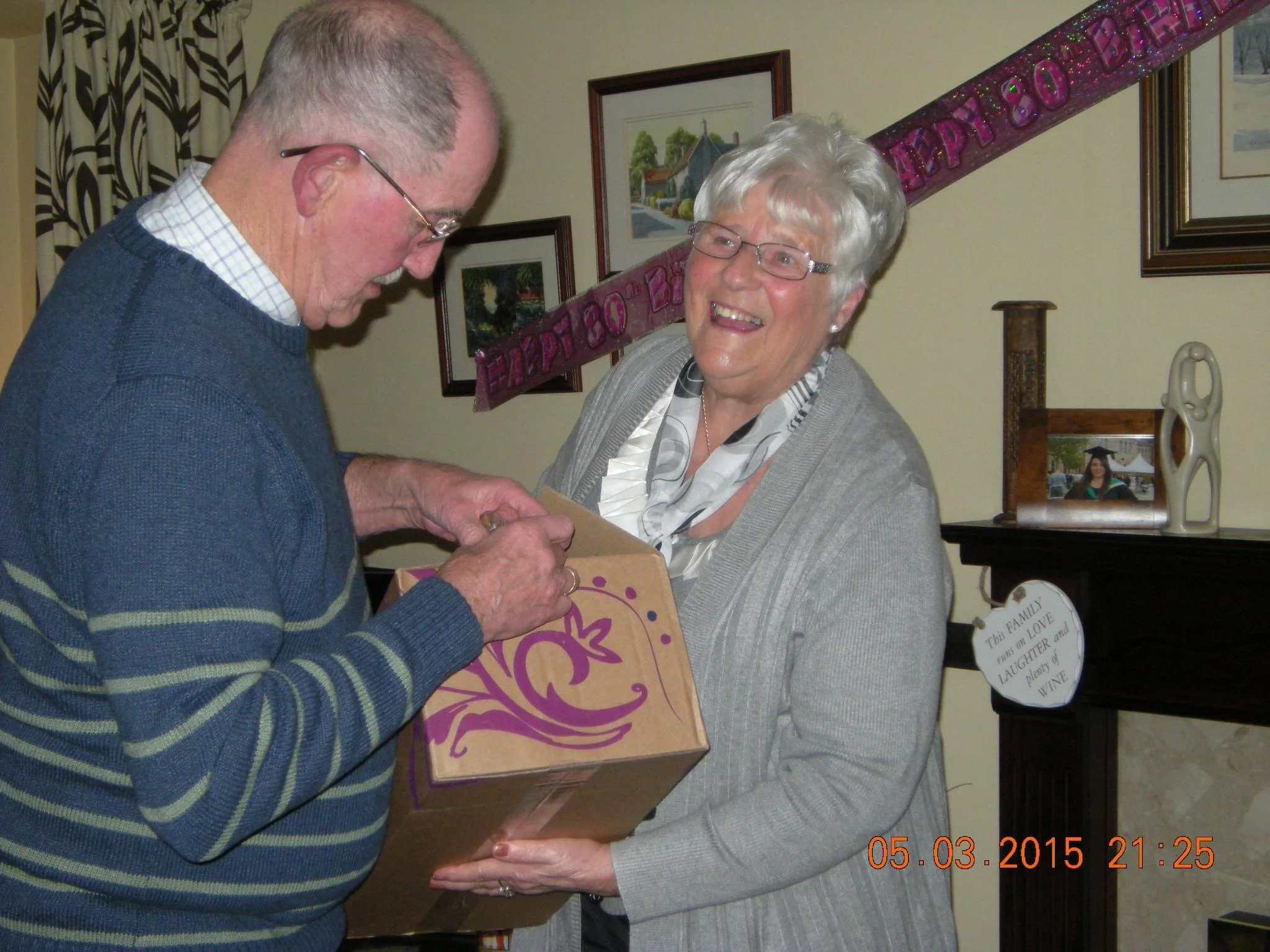 An elderly woman with gray hair and glasses is smiling and receiving a gift box from an elderly man with glasses. They are standing in a decorated room with family photos and a banner in the background, celebrating a special occasion.