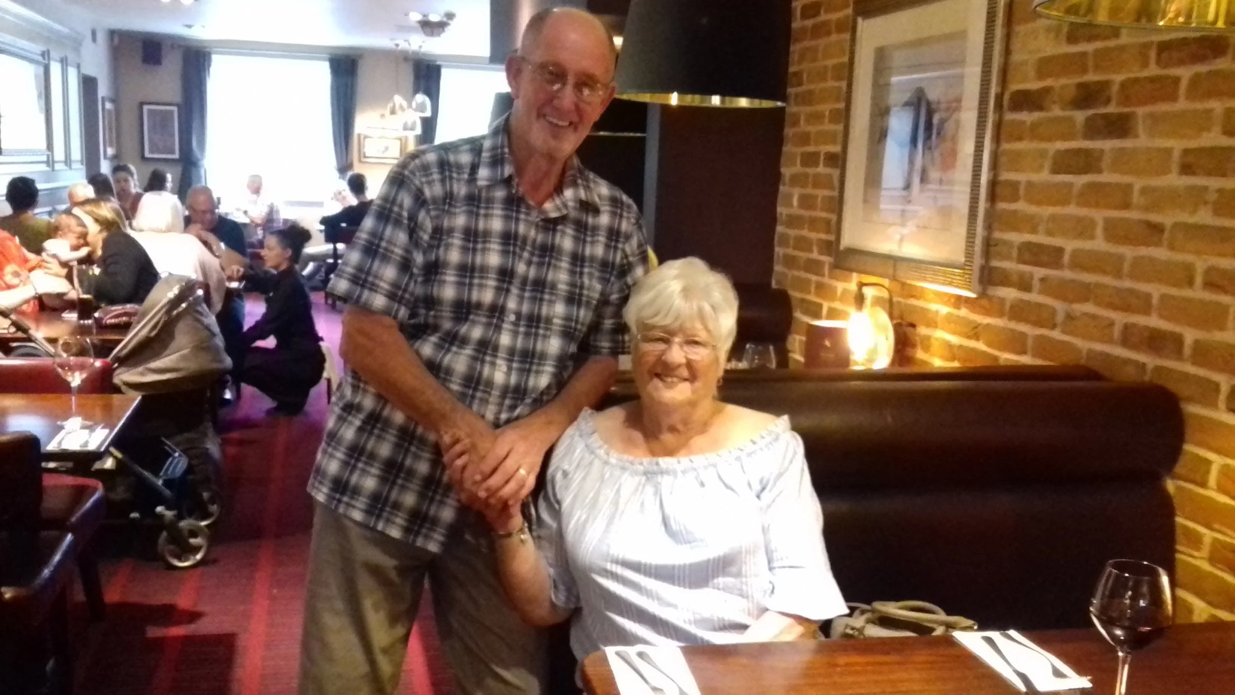 A smiling elderly woman sitting at a restaurant table with a man standing beside her, holding her hand. The restaurant has brick walls, framed art, and warm lighting, with other diners in the background.