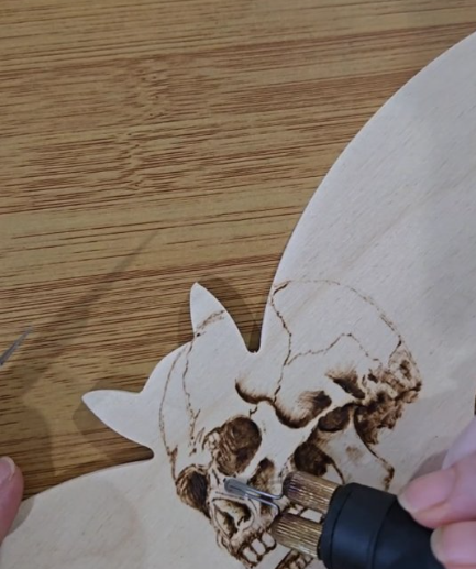 A person using a wood burner tool to burn a skull and spider web design onto a piece of light-colored wood, with another piece of wood in the shape of a cat's head.