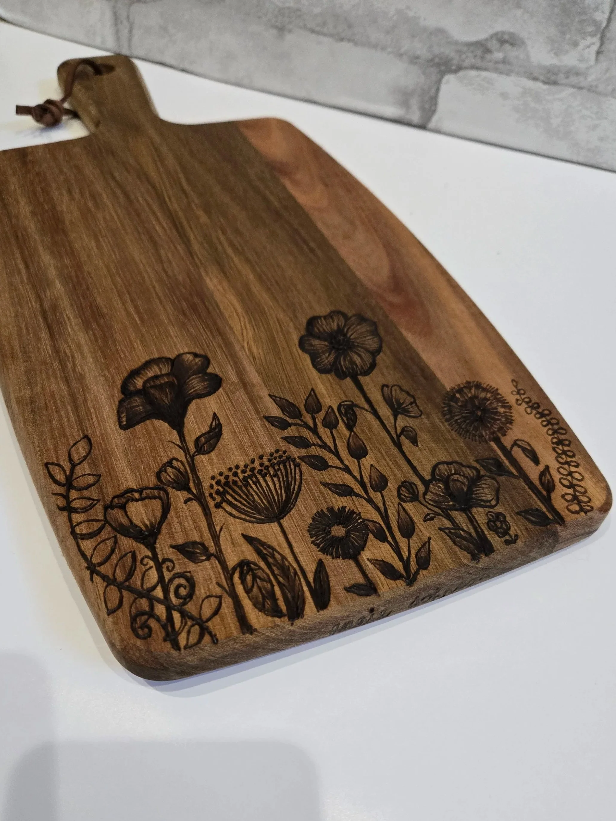 A wooden cutting board with flower illustrations engraved on it, placed on a white surface near a gray brick wall.