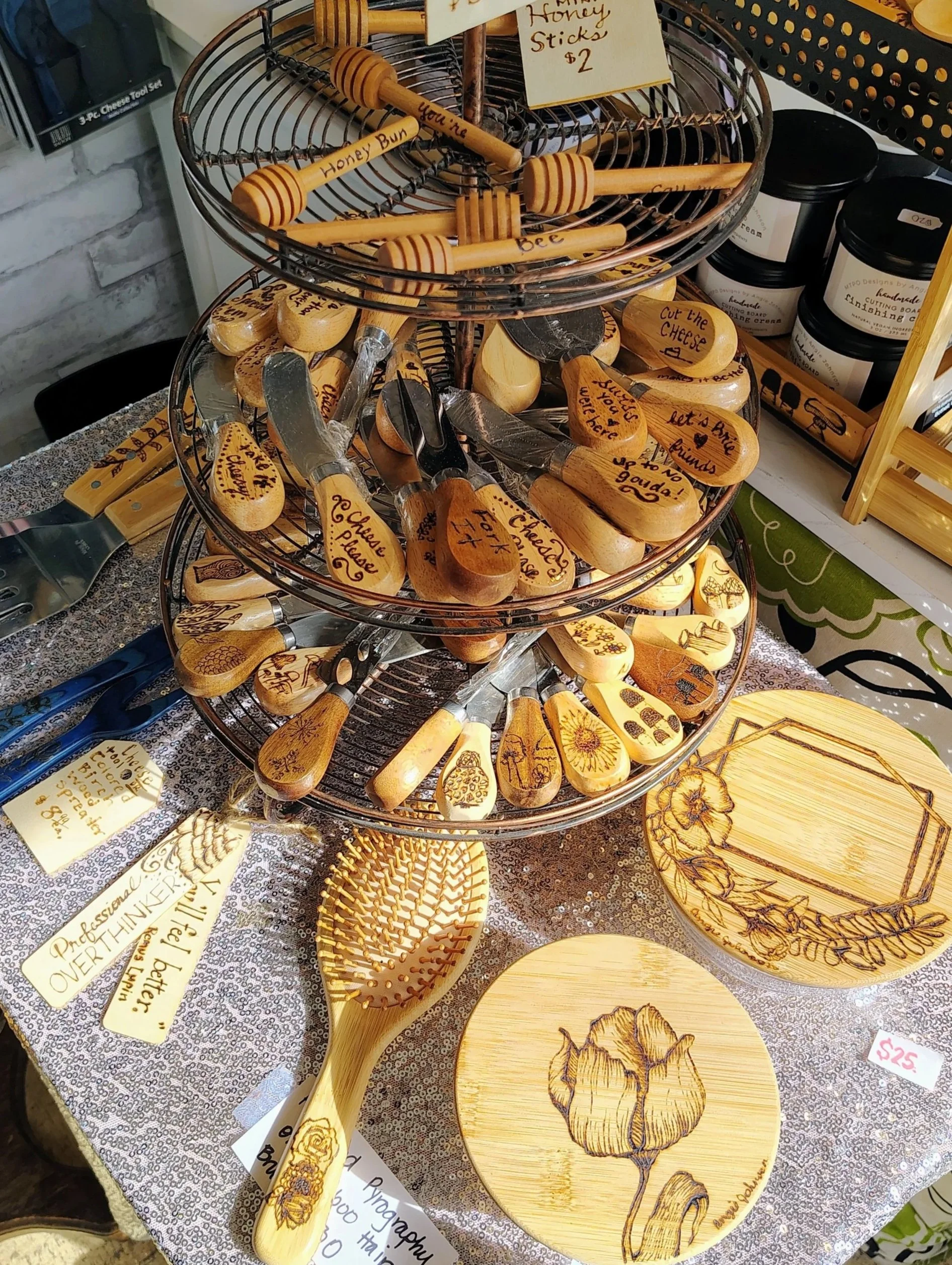 Wooden honey dipper with engraved text, wooden brush with engraved text, and wooden plaques with engraved flower designs and inspirational messages, all displayed on a table with various other handcrafted wooden items and jars in the background.