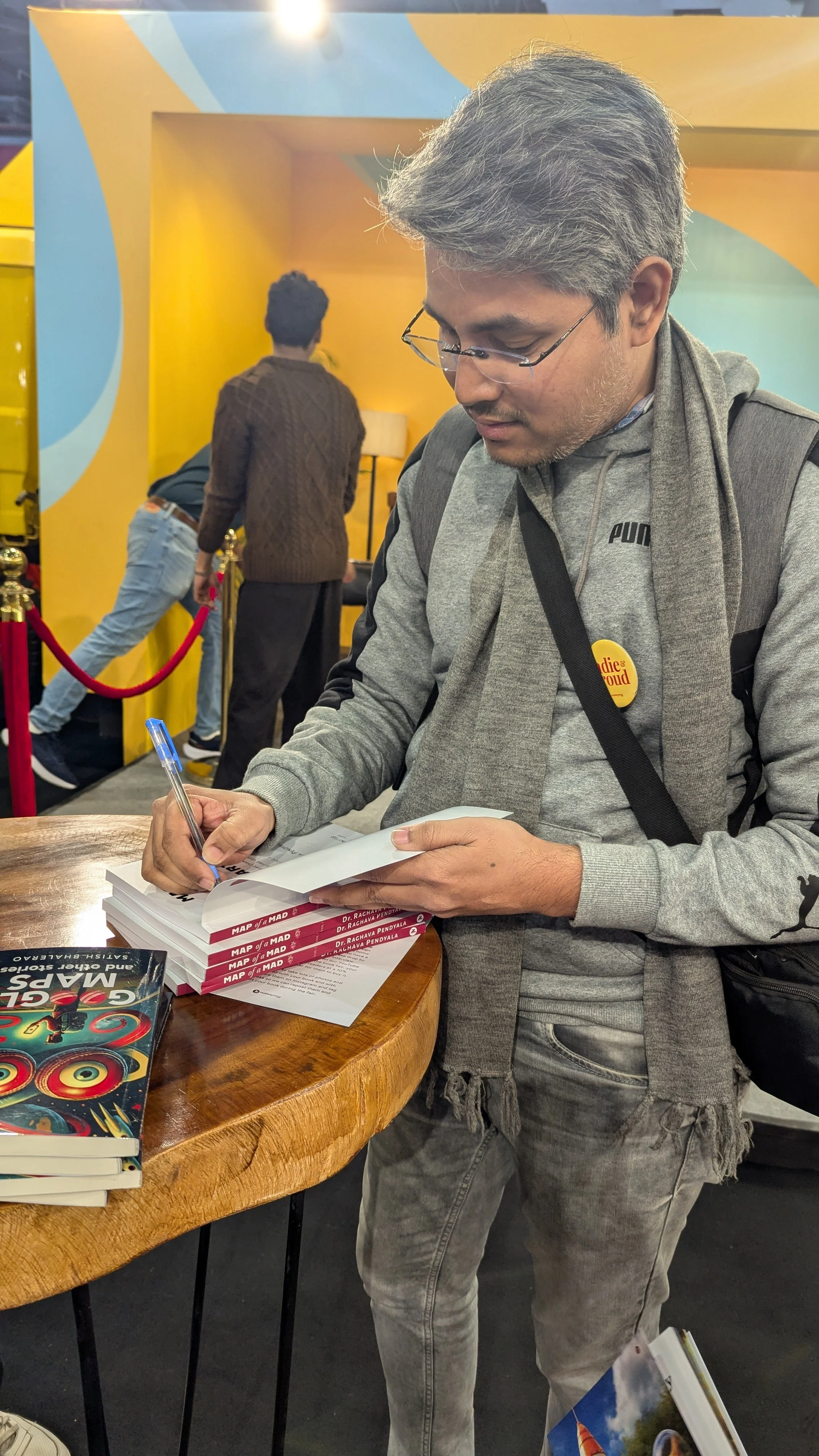 A man wearing glasses, a gray Puma hoodie, and a gray scarf is signing a book on a wooden table in a colorful indoor setting. There are several books stacked on the table, and a few people are visible in the background.