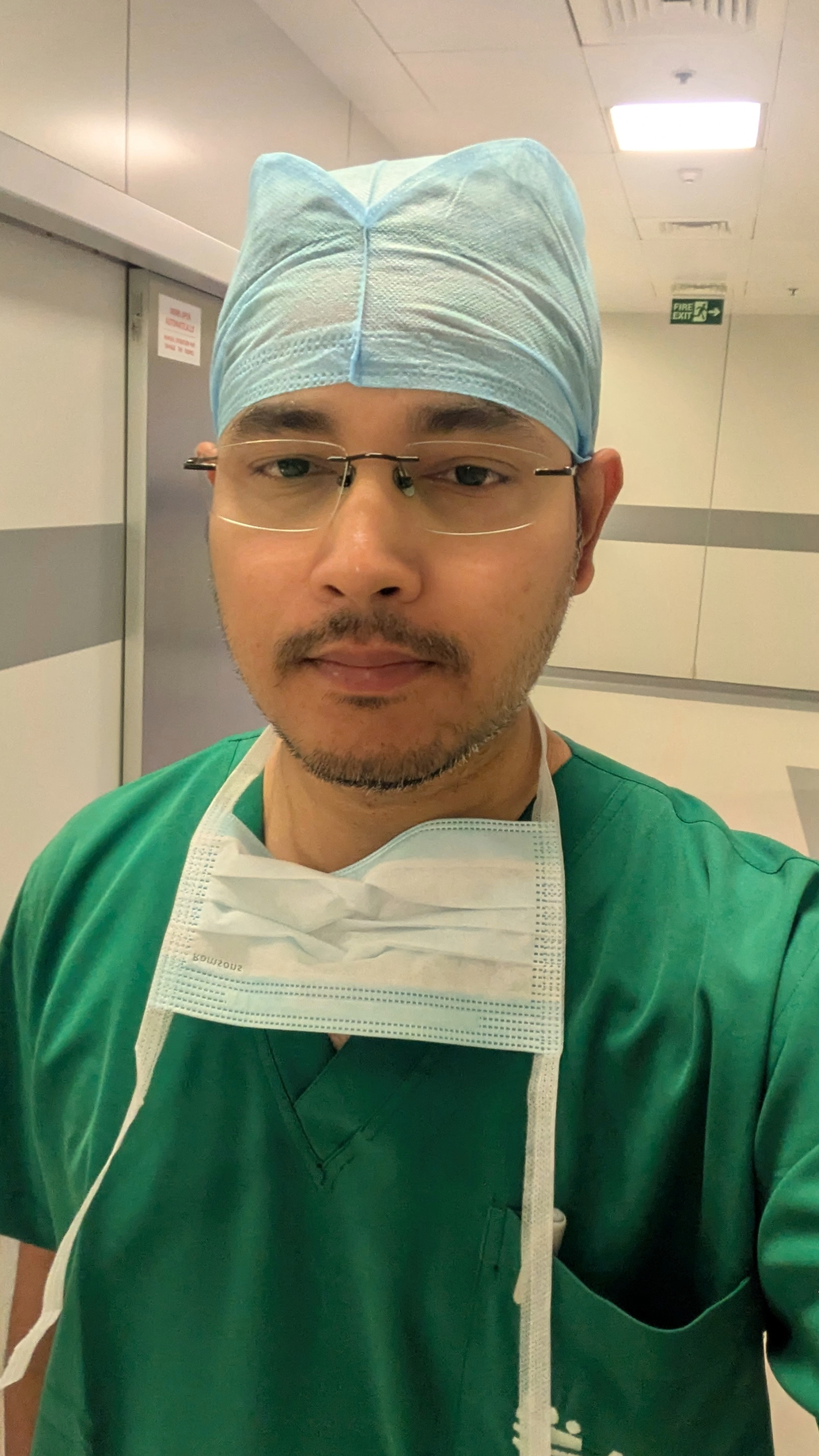 A male healthcare professional wearing green scrubs, a blue surgical cap, and a face mask hanging around his neck, taking a selfie in a hospital corridor.