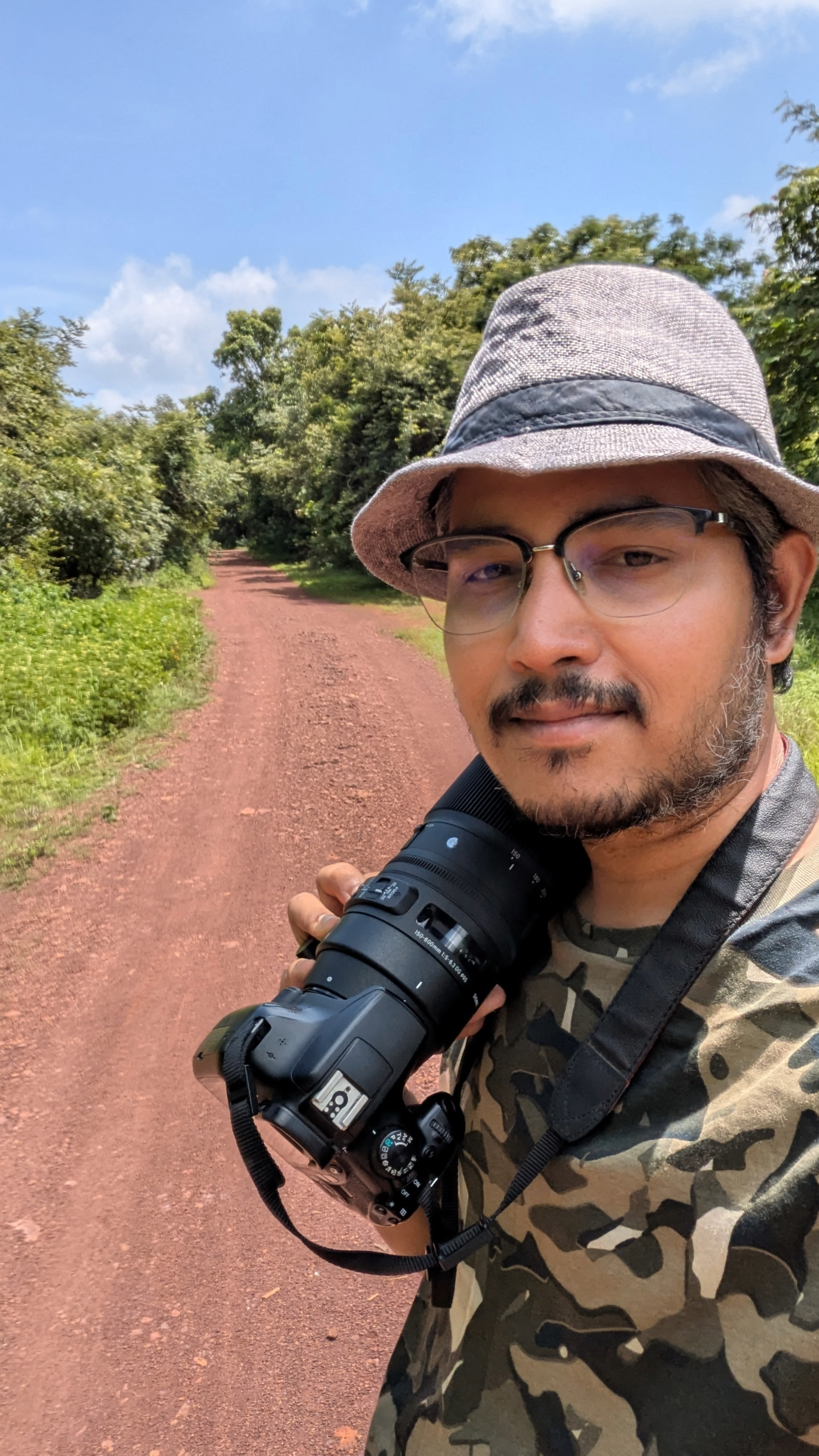A man wearing a gray hat, glasses, and a camouflage shirt is holding a camera on a red dirt path surrounded by green trees and blue sky.