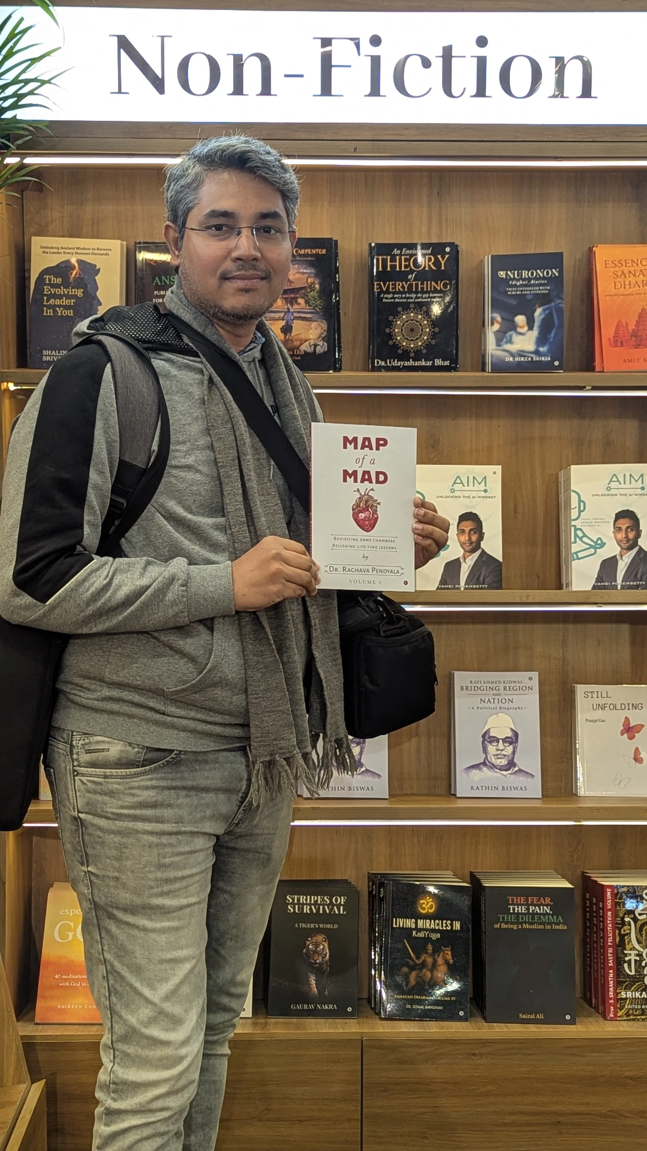 A man standing in front of a bookshelf filled with nonfiction books, holding a book titled "Map of a Mad Heart" by Dr. Raghava Pendyala, in a store or library.