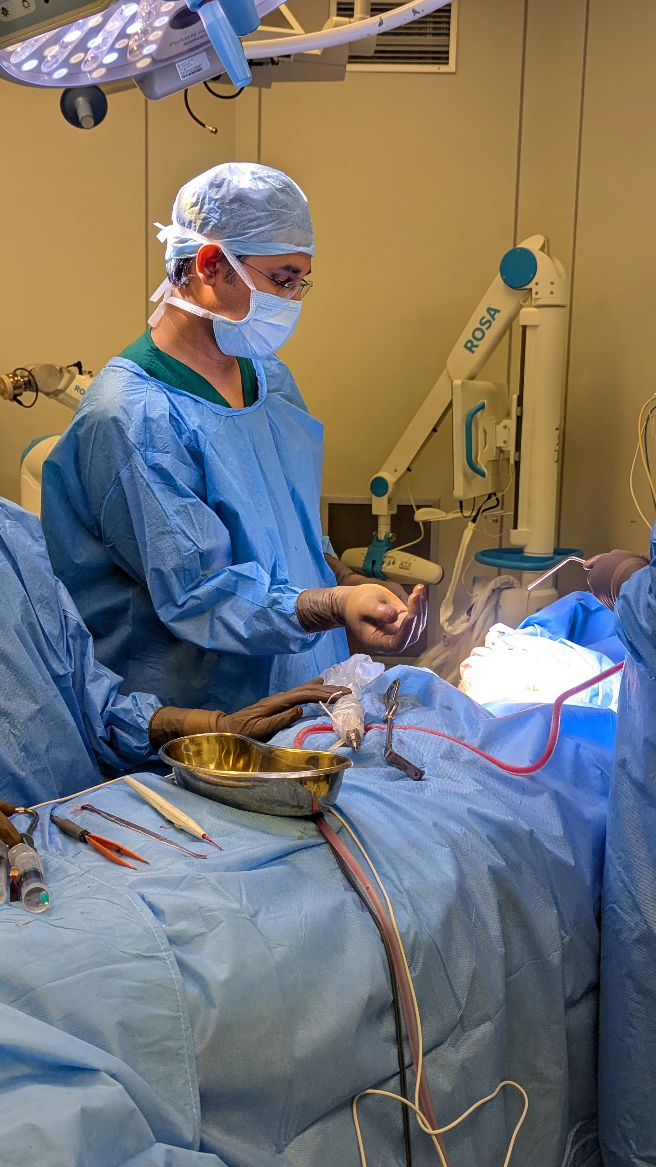 Surgeon in operating room performing surgery, wearing surgical gown, mask, and gloves, with medical equipment and surgical tools around.