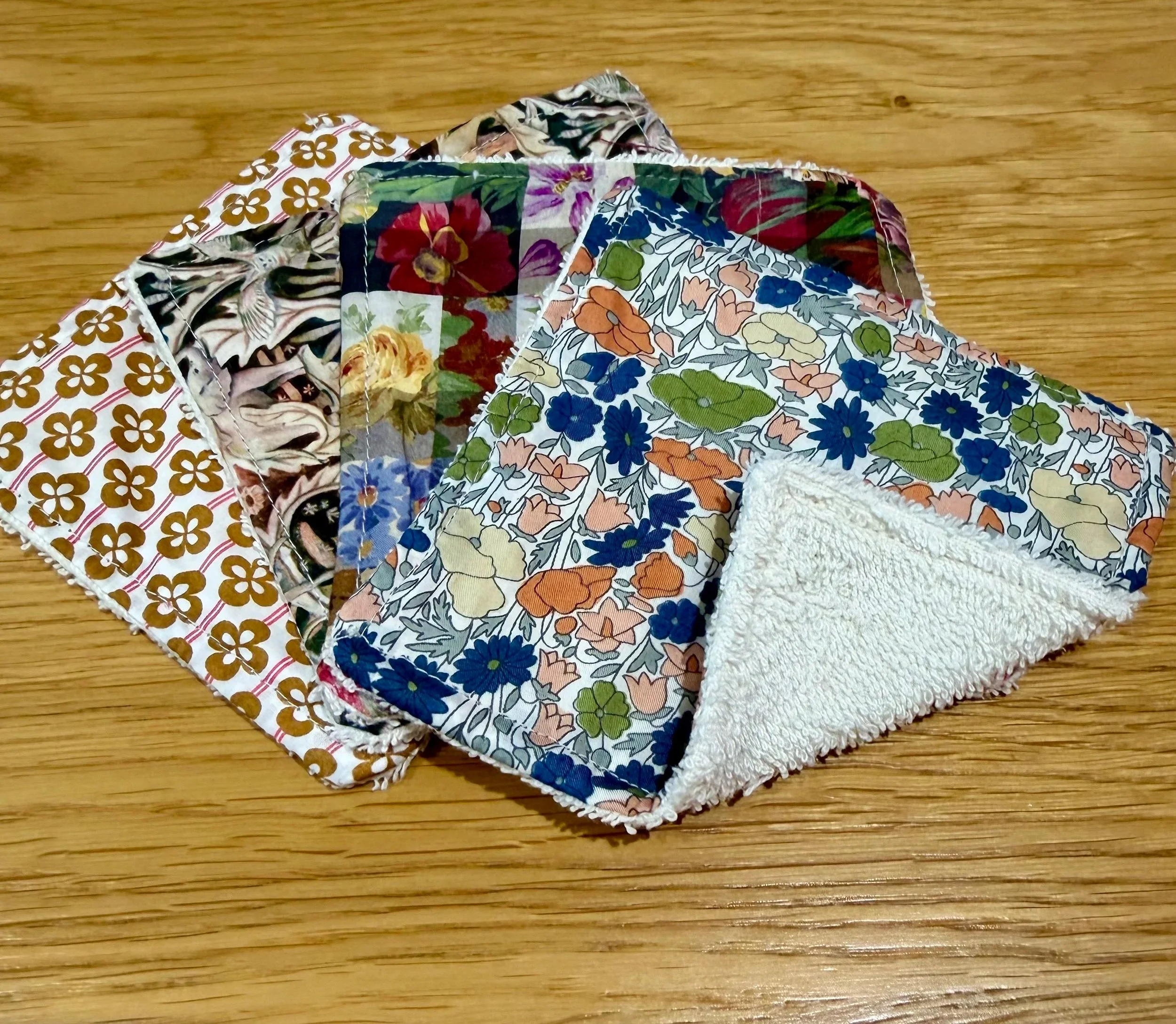 Reusable cleansing cloths arranged on a wooden surface, showing colourful Liberty floral cotton fronts and cream towelling backs.