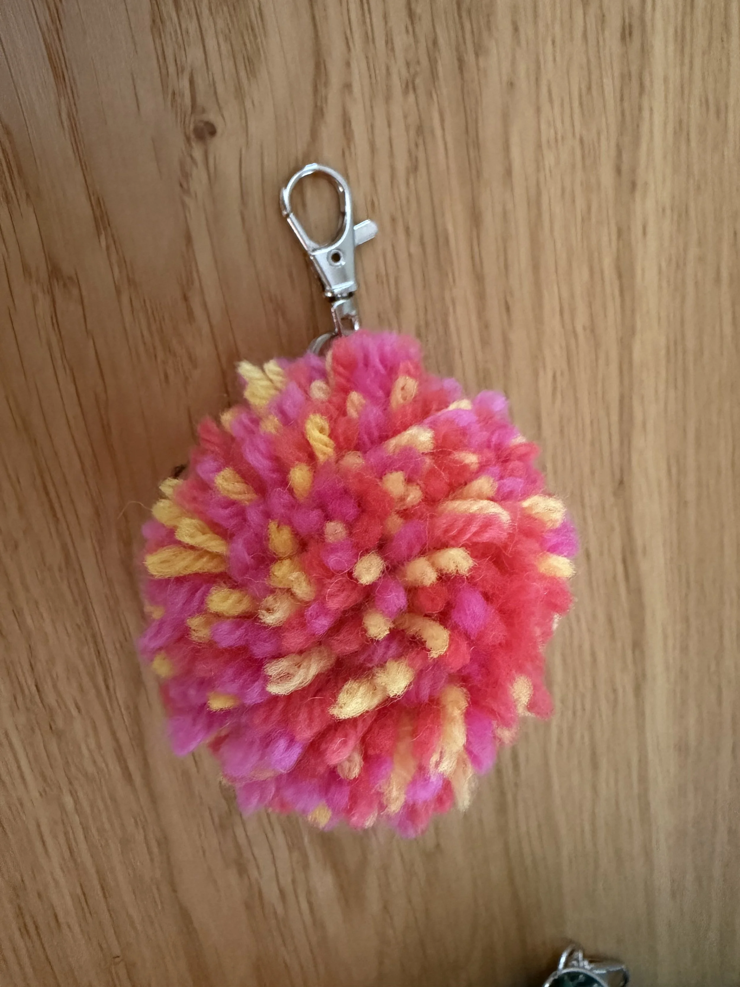 Handmade pink, coral and yellow wool pom pom key ring with a silver-coloured lobster clip on a wooden background.