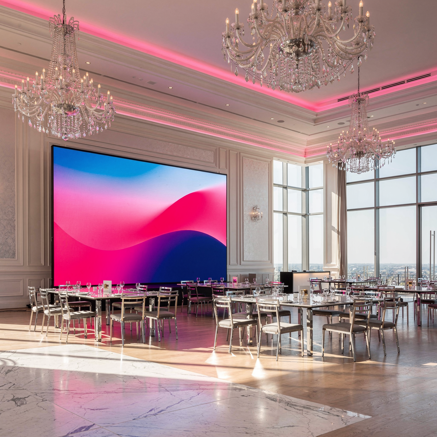 Eleganter festlicher Saal mit Kronleuchtern, großen Fenstern, runder Tafel und großem Bildschirm mit buntem abstraktem Hintergrund, für eine Veranstaltung vorbereitet.