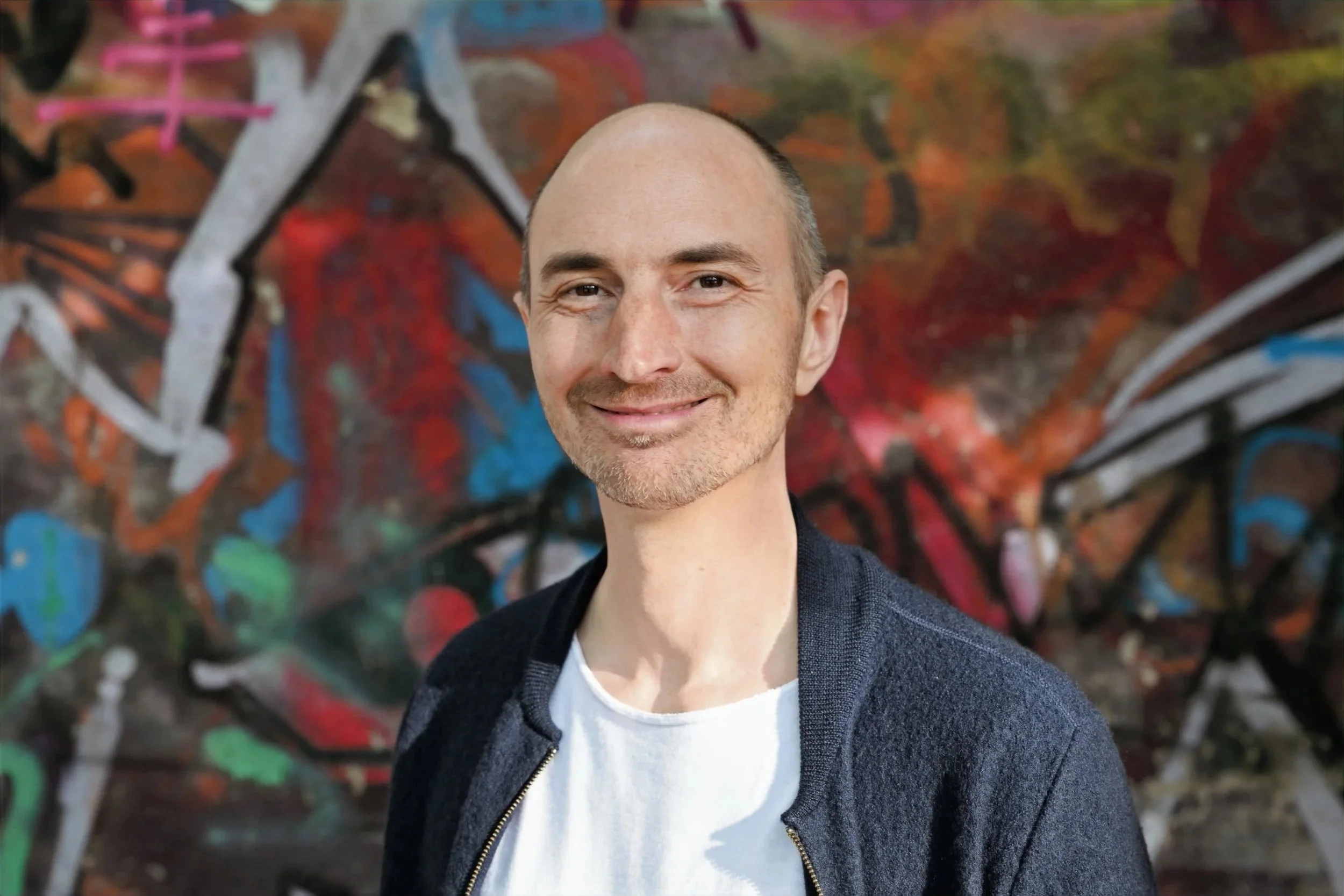 Ein Mann mit glattem Kopf und leichtem Bart smiling vor einem Graffiti-Hintergrund.