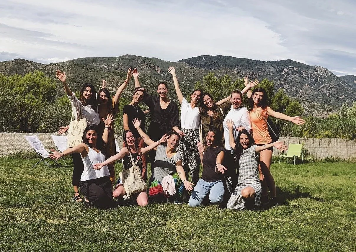 Grupo de diez y ocho jóvenes, sonriendo y levantando los brazos, en un ambiente al aire libre con montañas y cielo nublado al fondo.