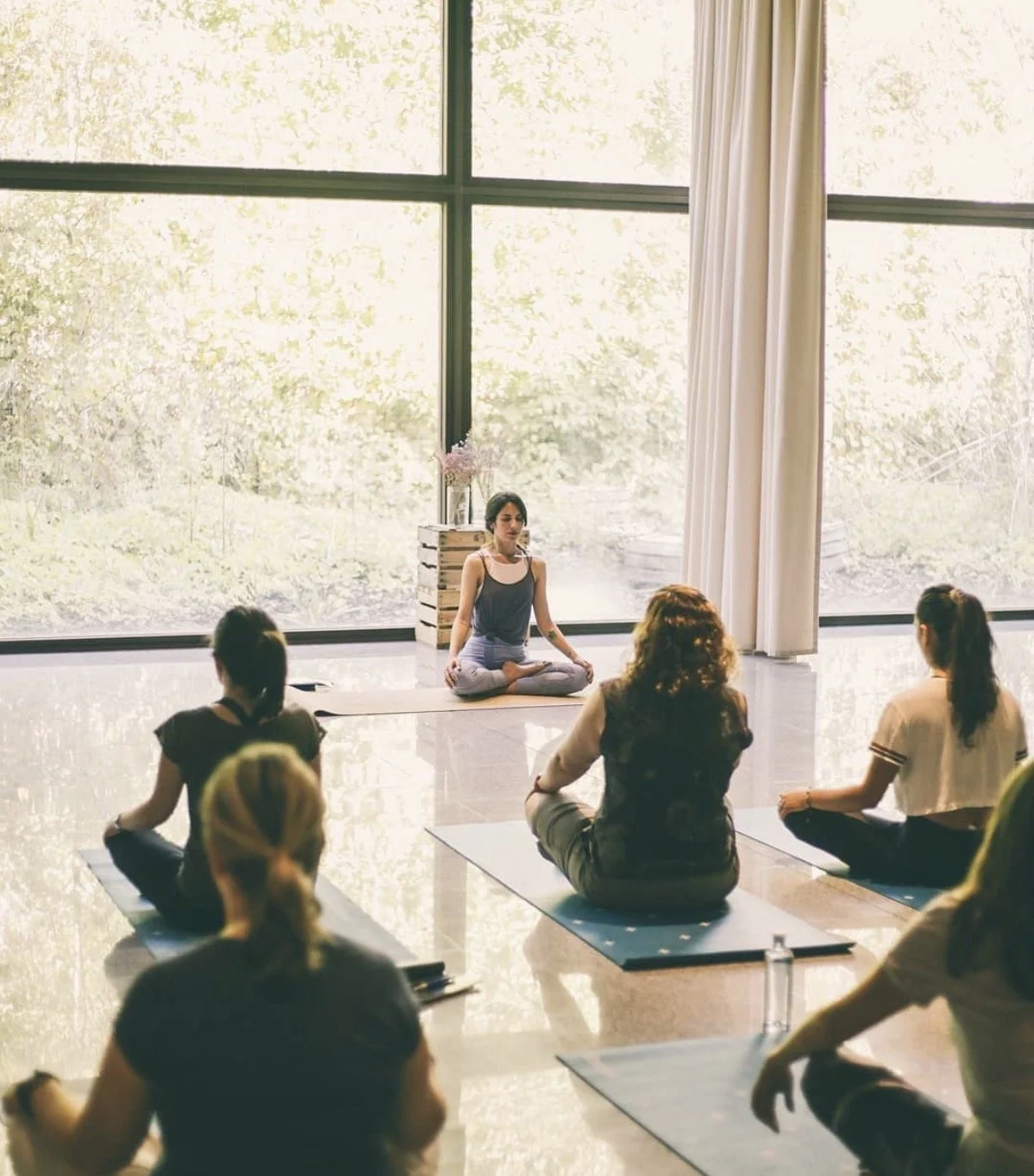 Mujer haciendo meditación en una sesi f3n de yoga o mindfulness, rodeada de varias personas sentadas en postura de loto en una habitaci f3n luminosa con paredes de cristal que muestran un  e1rbol y un paisaje natural.