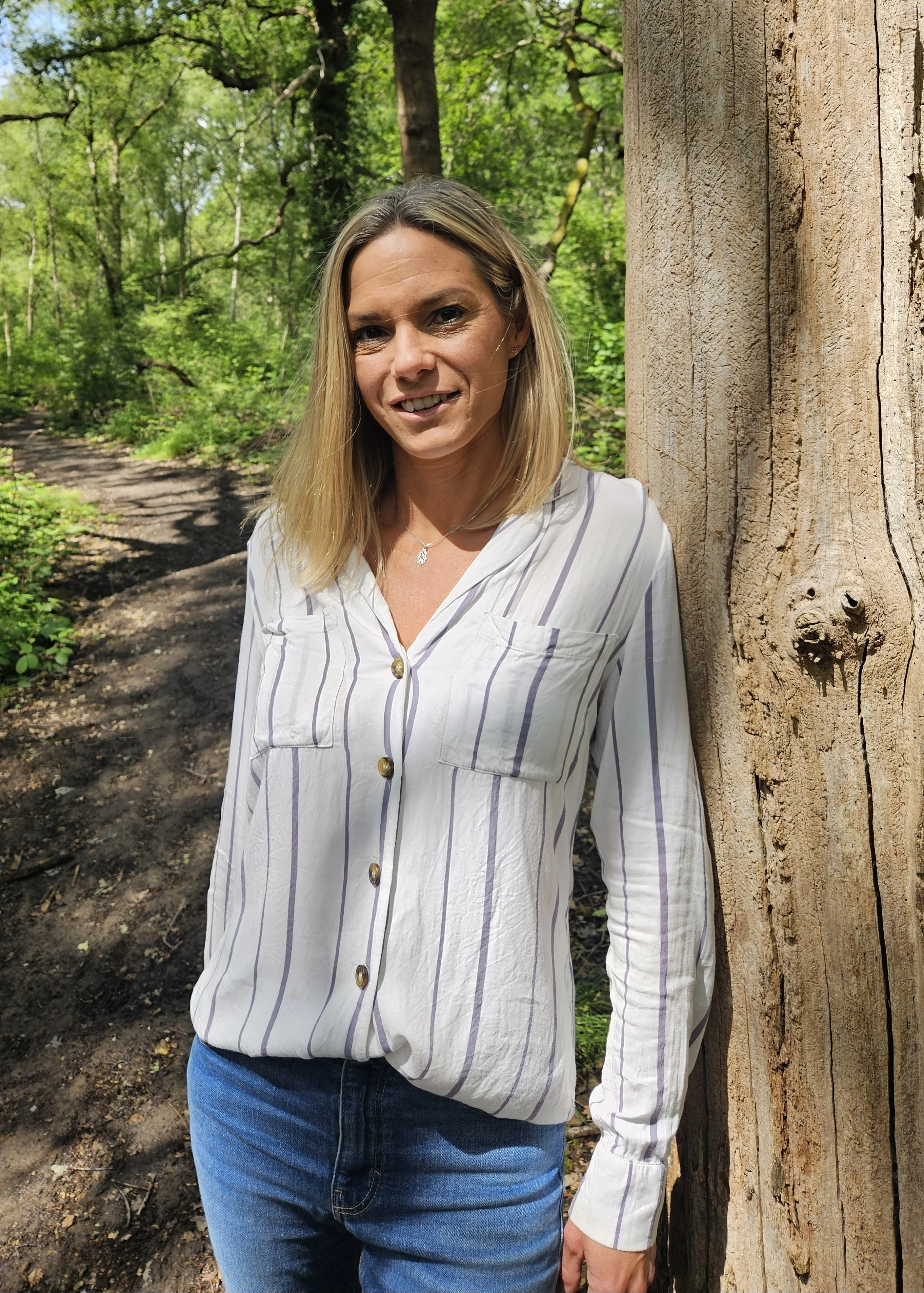 A woman with blonde hair standing outdoors next to a large tree trunk in a forested area with green foliage, wearing a white striped button-up shirt and blue jeans.