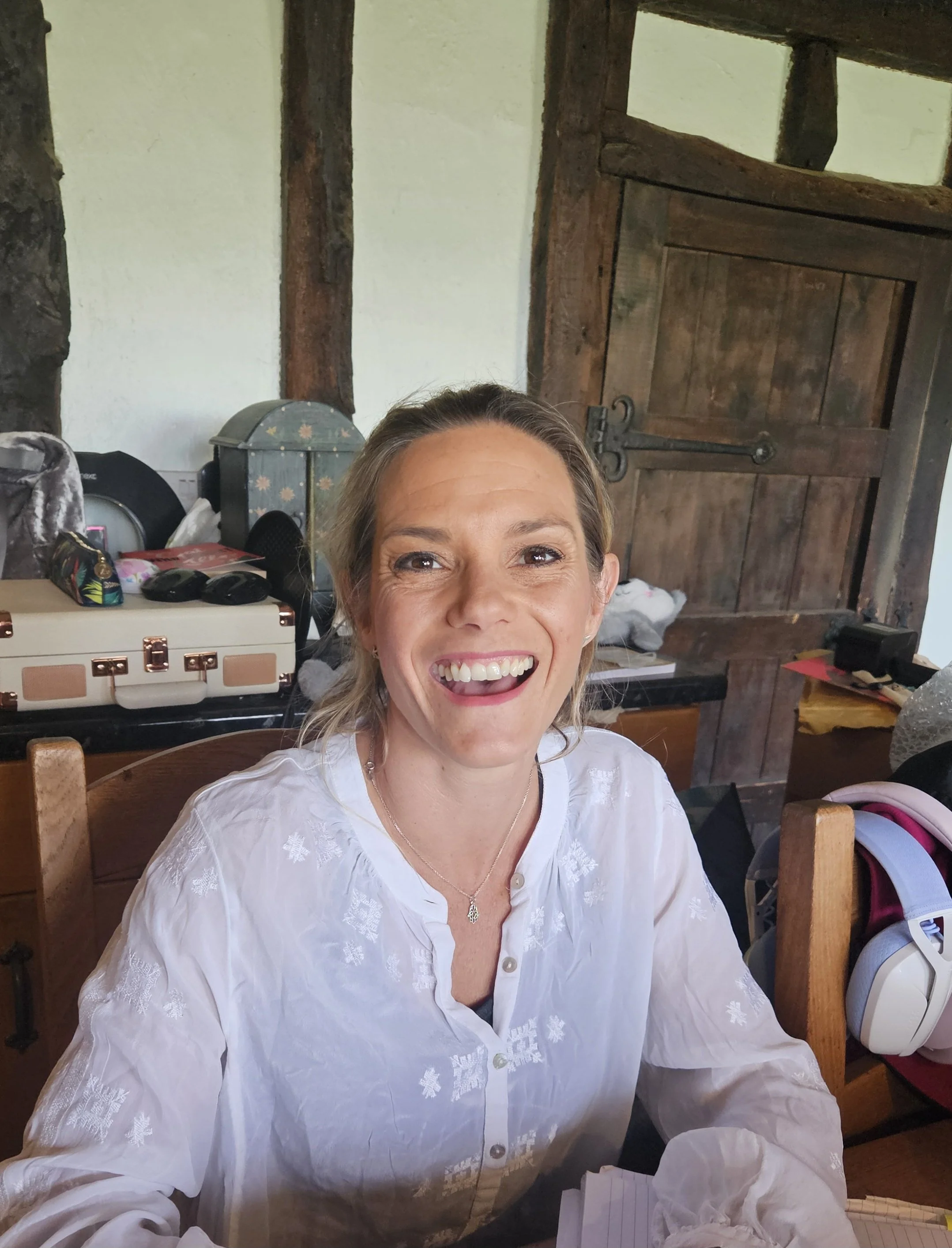 A smiling woman with blonde hair wearing a white embroidered blouse, sitting indoors with a rustic wooden door and various items in the background.