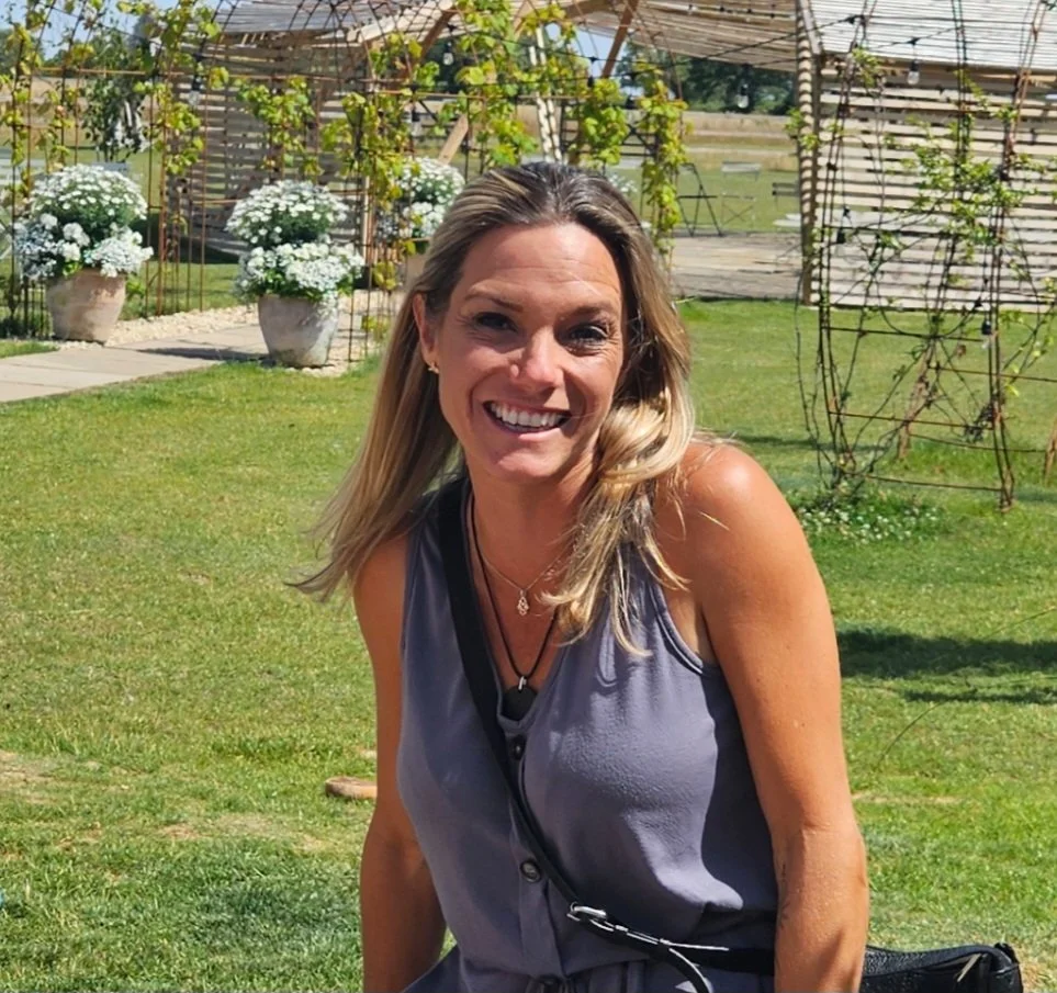 A woman smiling outdoors in a garden with white flowers and trellises in the background.