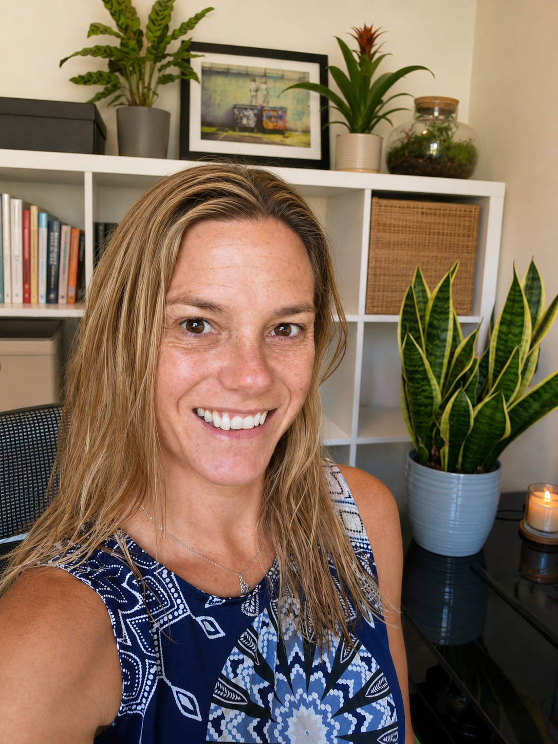 A woman with long, wet blonde hair smiling in a room with a white bookshelf, houseplants, framed photo, and a lit candle in the background.