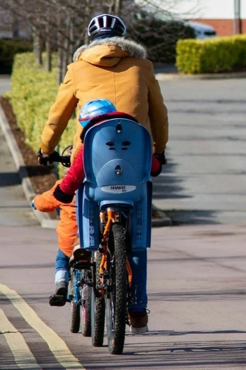An adult and a child riding bikes together on a street. The child has a blue helmet and is seated in a blue child bike seat, while the adult wears a black helmet and a yellow jacket. The scene is outdoors with trees and bushes along the sidewalk.