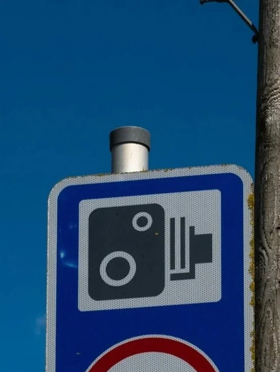 Close-up of a blue road sign indicating a speed camera, mounted on a wooden pole with a clear blue sky in the background. Wood street, Walthamstow, London.