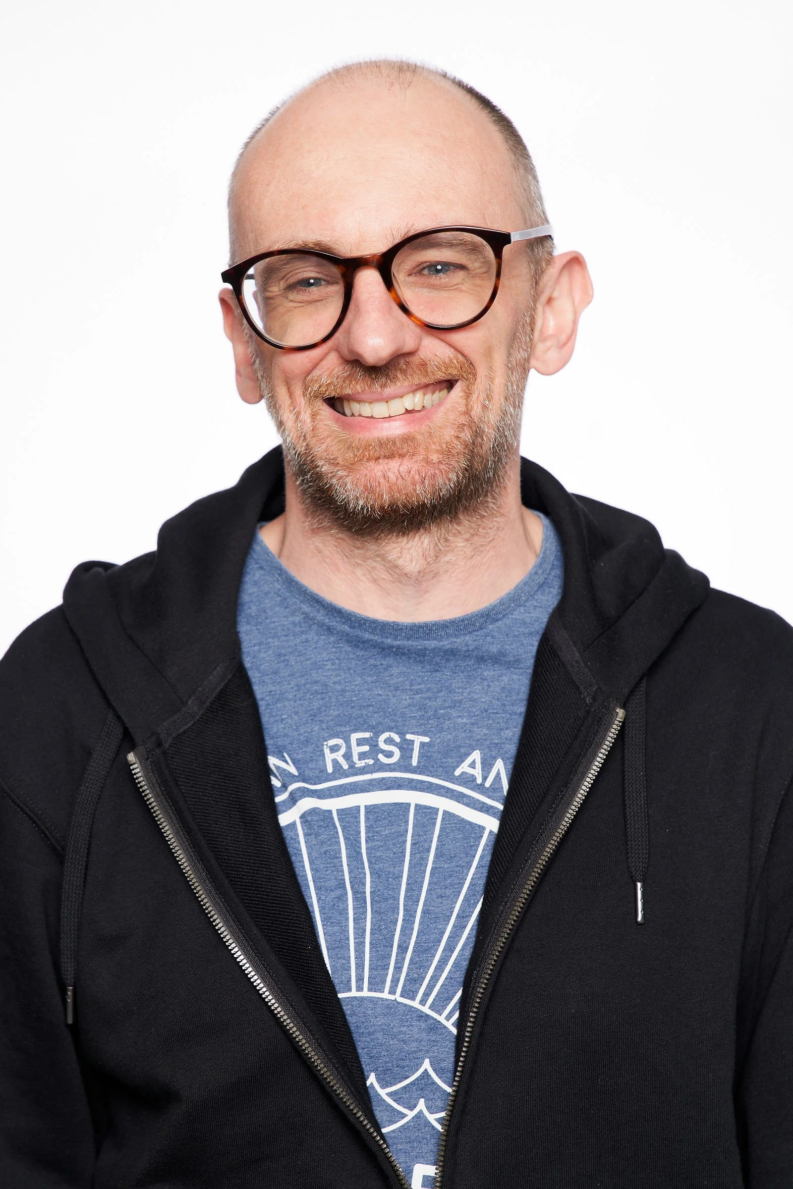 A smiling man with glasses and a beard, wearing a black hoodie and a blue T-shirt featuring a white design, against a plain white background.