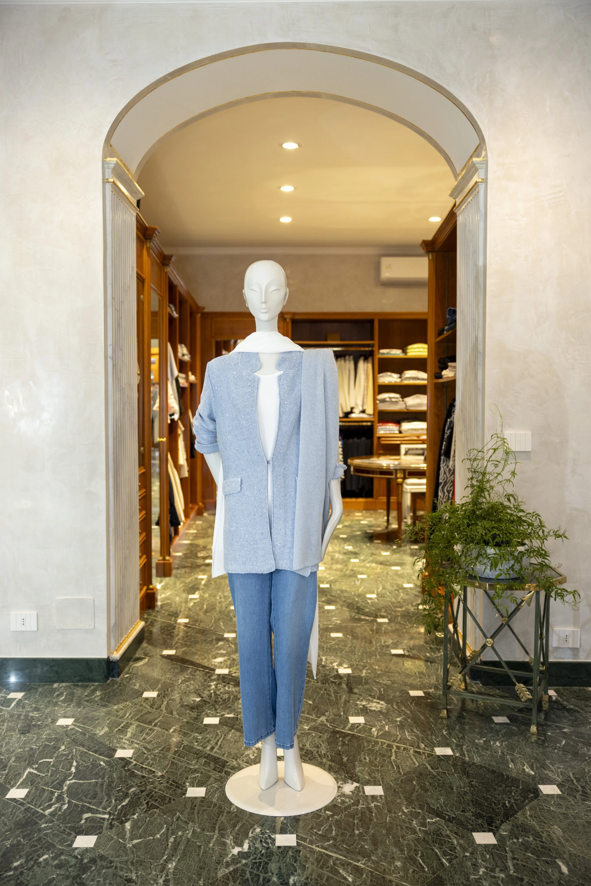 Statua di un manichino femminile con abbigliamento casual, pantaloni blu e giacca grigia, in un negozio di abbigliamento con scaffali di vestiti sullo sfondo e pavimento in marmo.