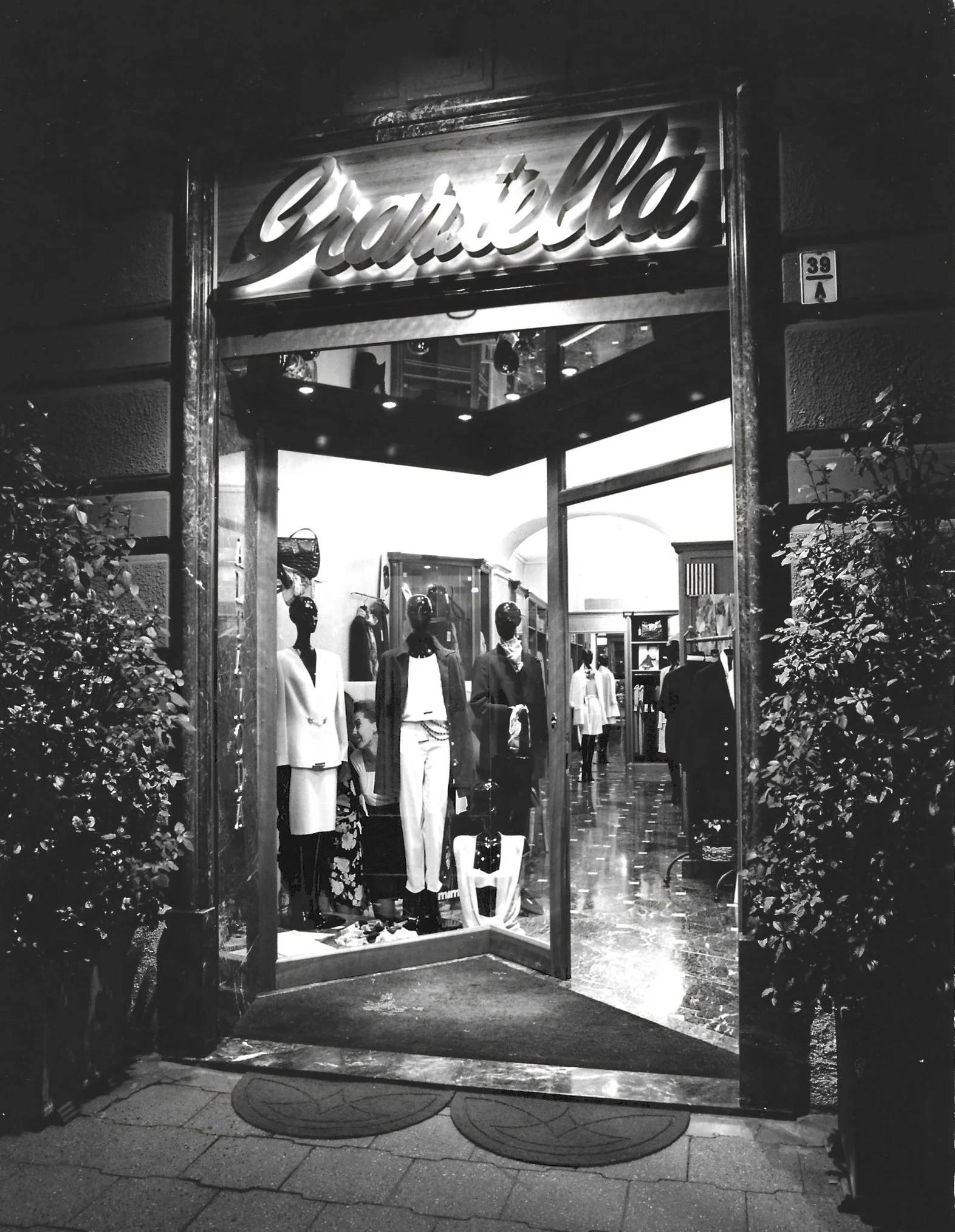 Una vetrina di un negozio di abbigliamento con la porta aperta, illuminata dall’interno, con manichini vestiti e una scatola con cappelli o borse. La scena è in bianco e nero.