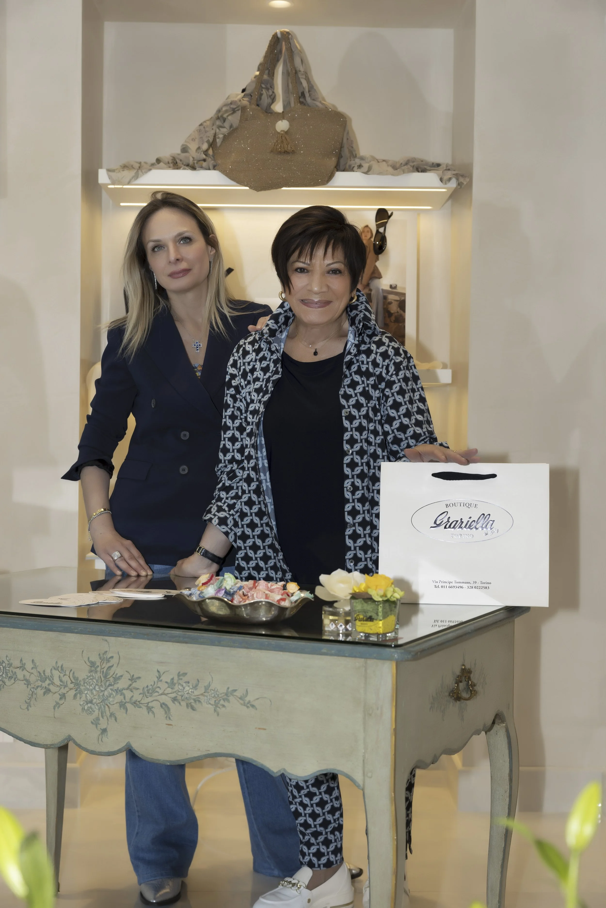 Due donne sorridenti davanti a un tavolo con fiori e un sacchetto di carta con il logo Graziella Boutique, in un ambiente elegante. Sulla mensola dietro di loro, una borsa di stoffa con sciarpa attorcigliata.