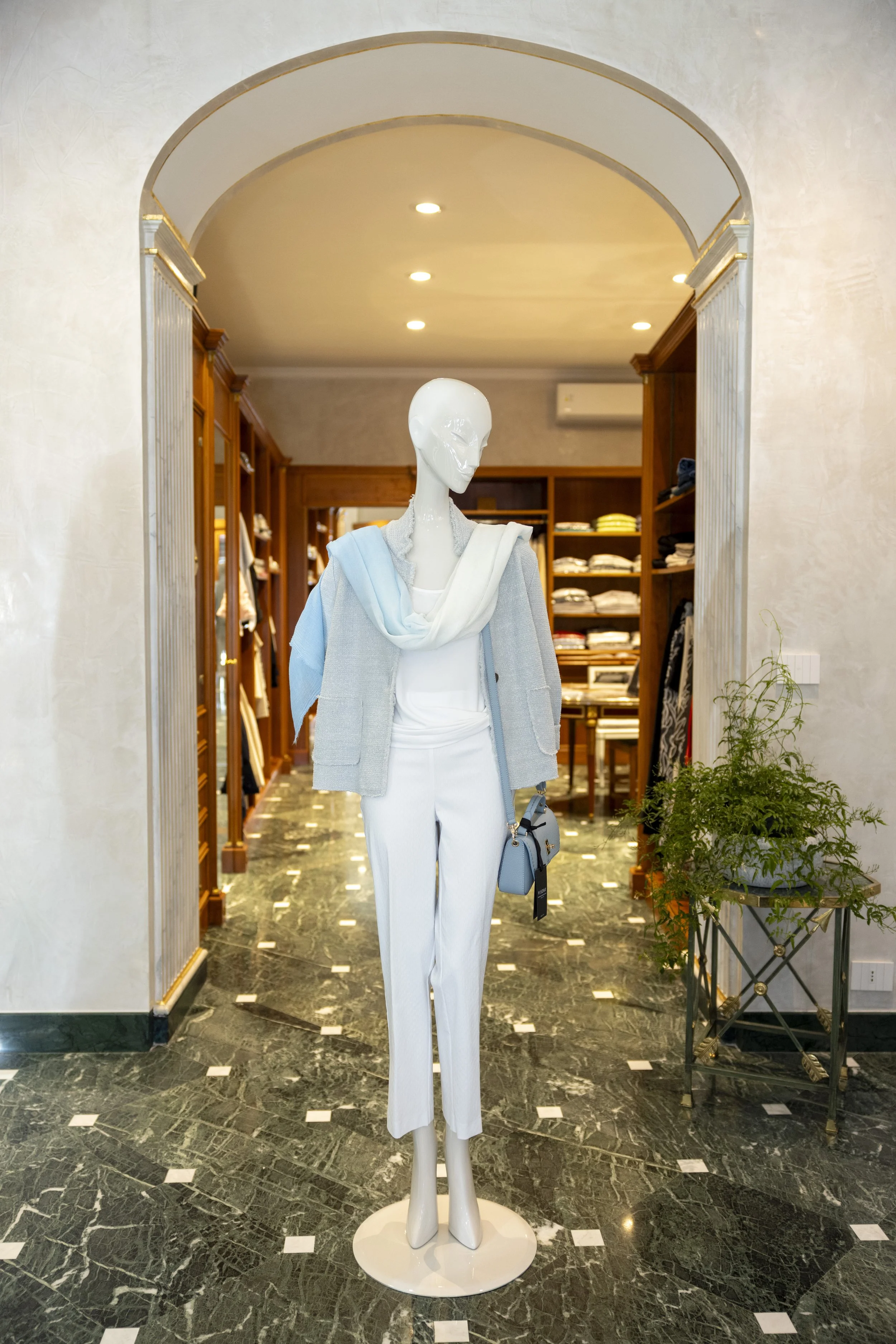 Manichino vestito con abito bianco, cappotto grigio chiaro, e sciarpa azzurra, in un negozio di abbigliamento con scaffali di vestiti e un pavimento in marmo verde scuro.