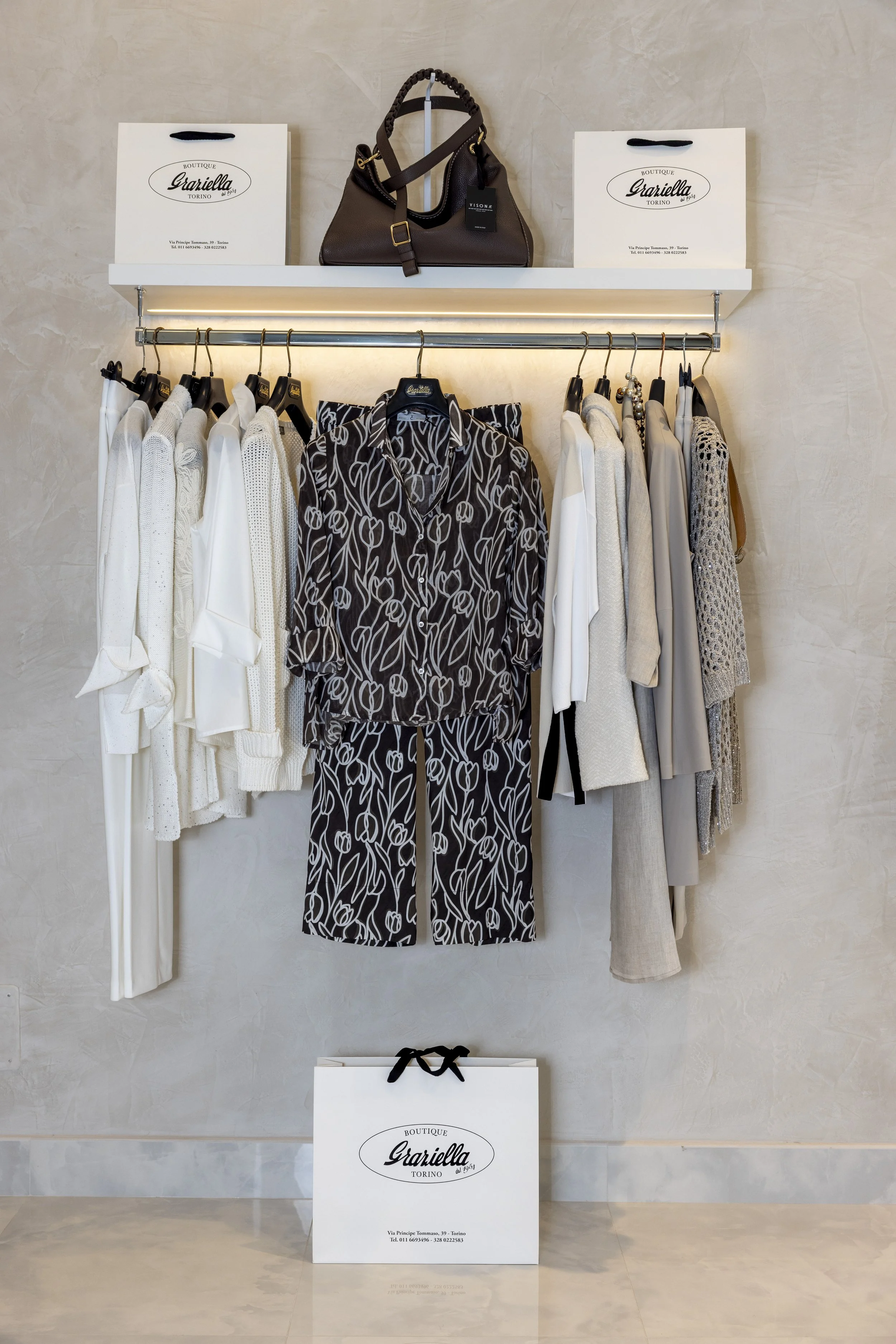 Esposizione di abiti e accessori moda in un negozio, con borse e capi di abbigliamento di vari stili e colori, presentati su una gruccia e una mensola.
