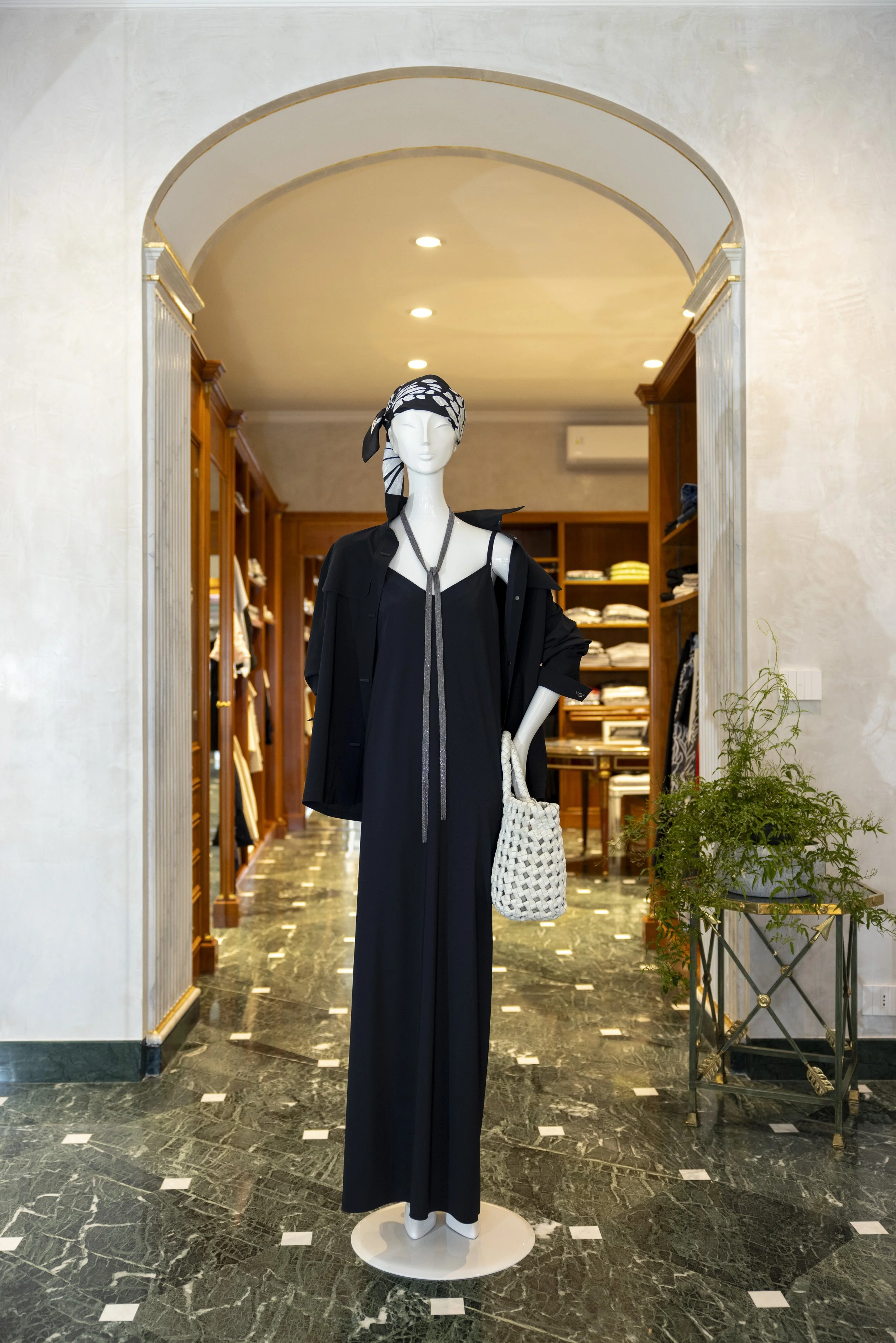 Manichino in abito nero lungo con sciarpa e borsa bianca a rete, decorato con bandana nera e bianca, in un negozio di moda interno con scaffali di vestiti e piante verdi a fianco.