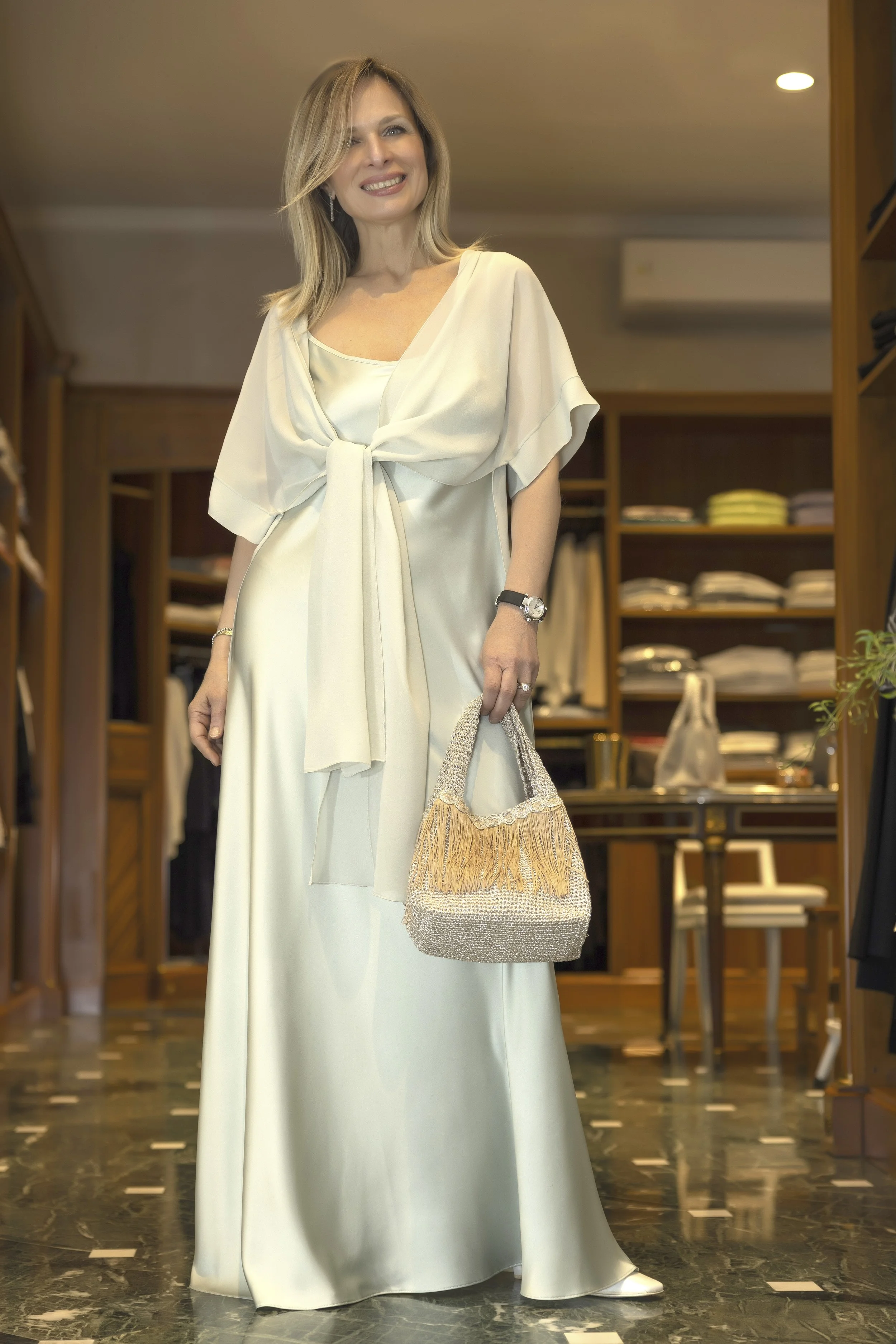 Donna sorridente in vestito lungo bianco con dettagli in seta, che tiene una borsa con frange in un negozio di abbigliamento.