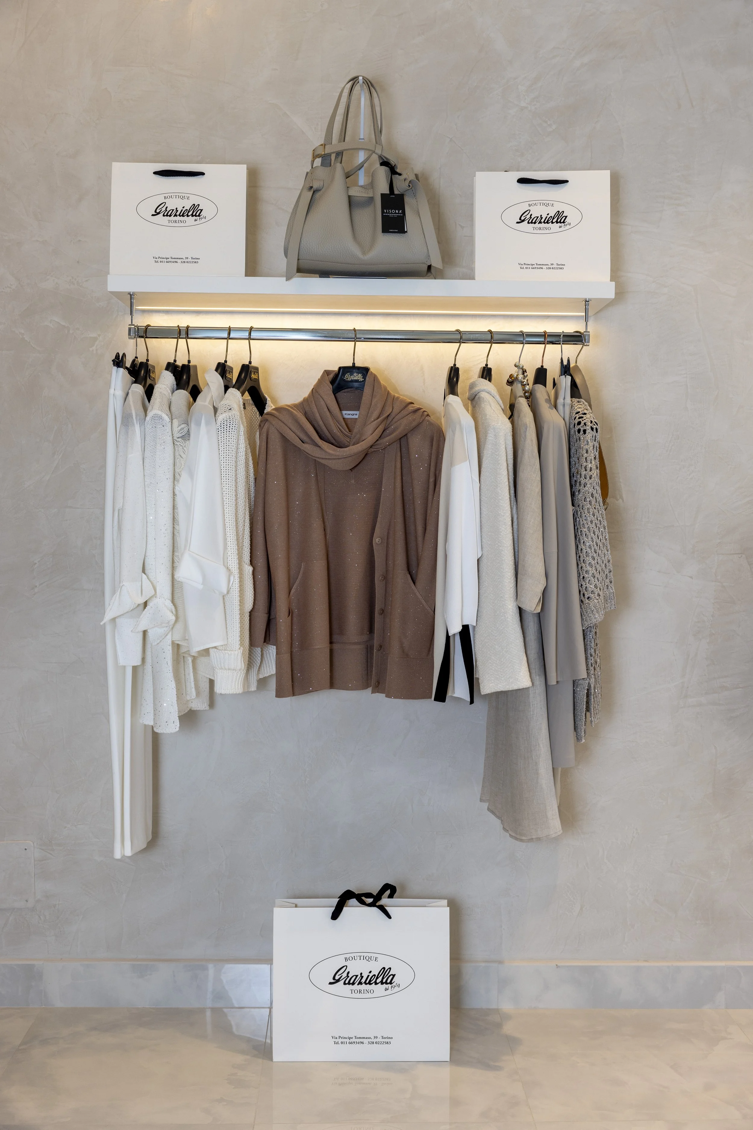 Stand di abbigliamento in un negozio con una rastrelliera di vestiti bianchi, beige e marroni, una borsa grigia sopra una mensola bianca e sacchetti di carta con il logo 'Garella'.