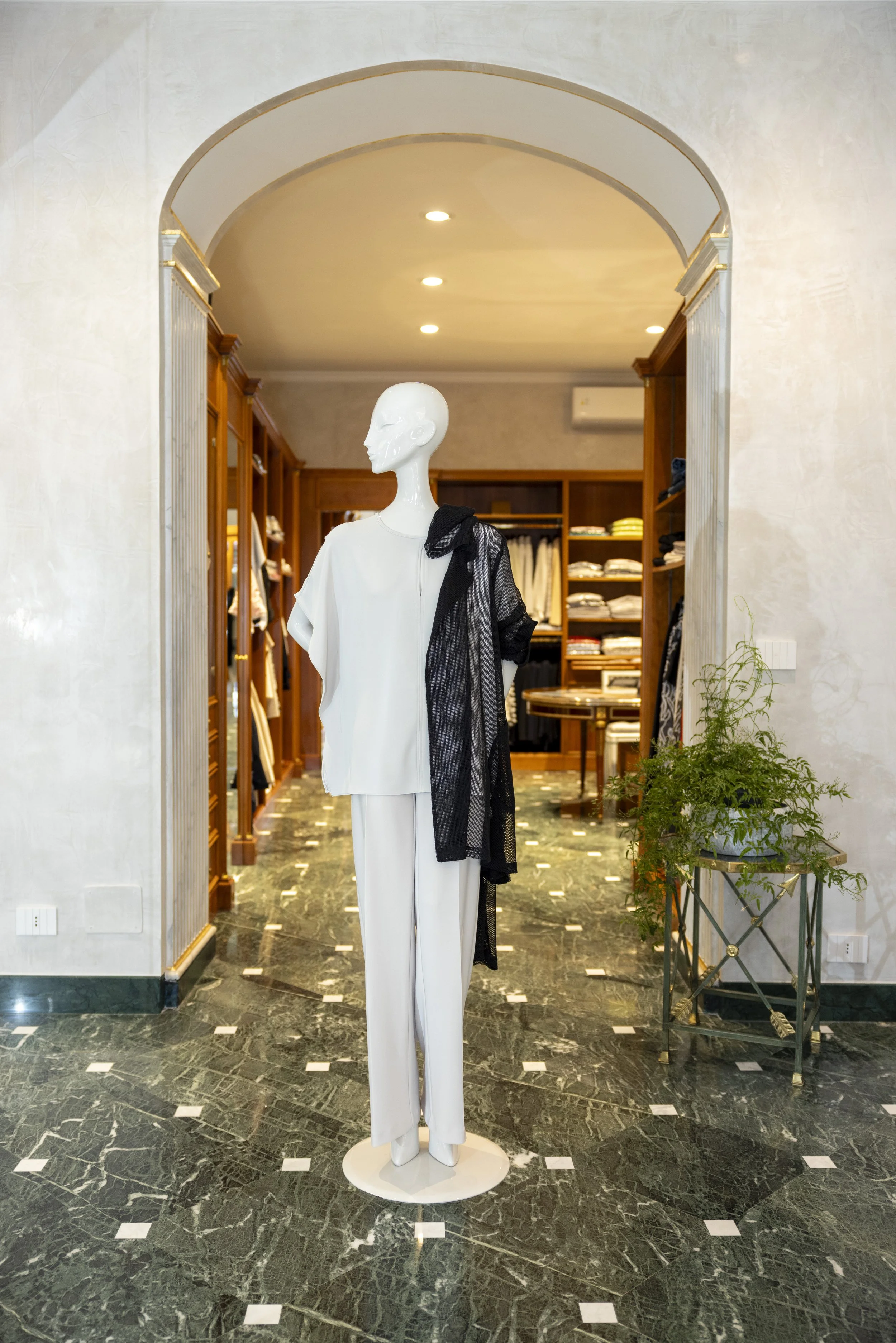 Manichino vestito bianco con nero in uno store di abbigliamento, pareti di marmo scuro, scaffali di legno con vestiti e armadio alle spalle, pianta verde a destra.