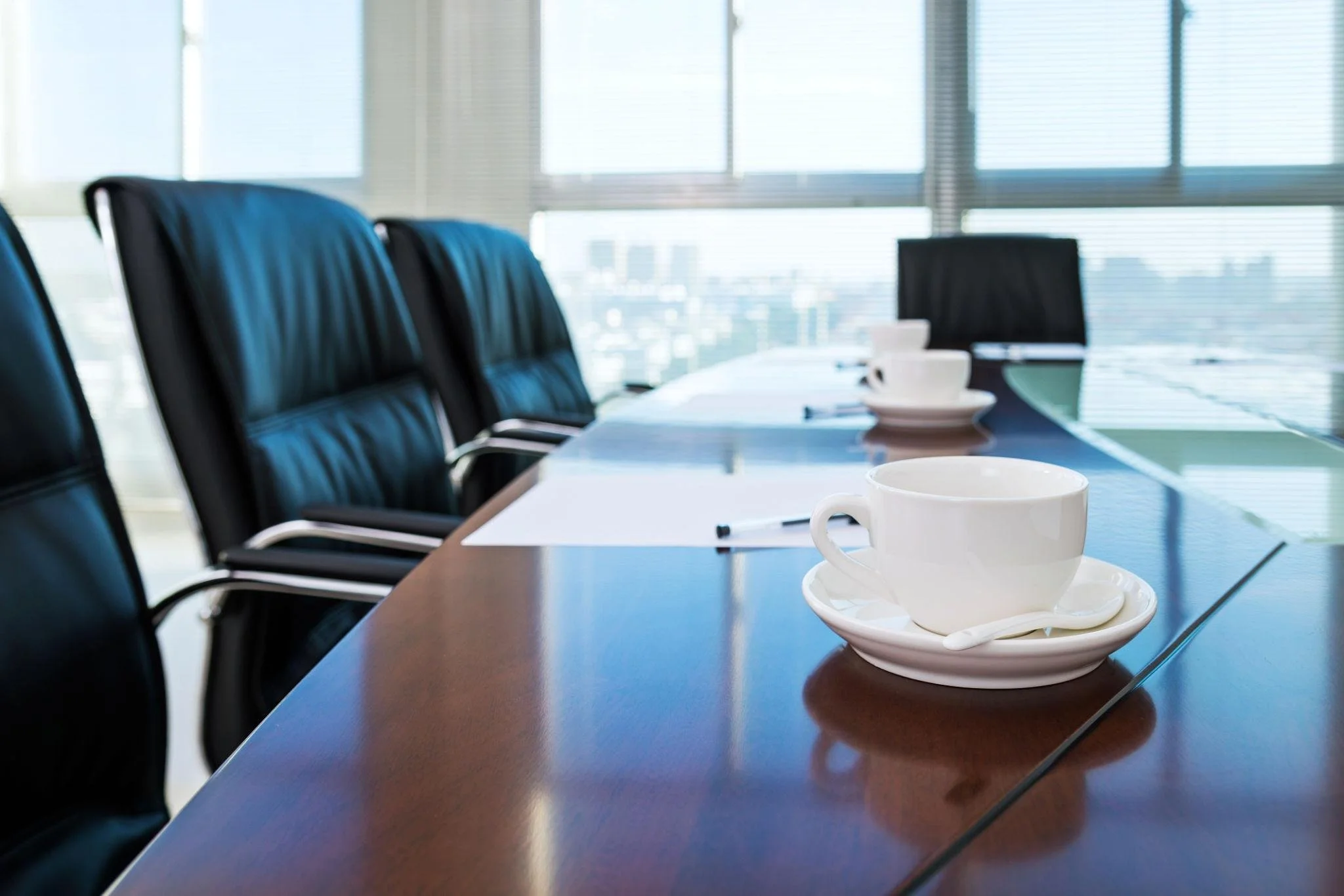 Conference room with white coffee cups on saucers and a marker on a polished wooden table in front of leather chairs and large windows.