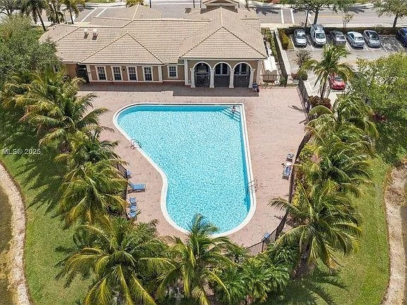 Aerial view of a backyard with a swimming pool surrounded by palm trees, a patio, and a house with a parking lot in the background.