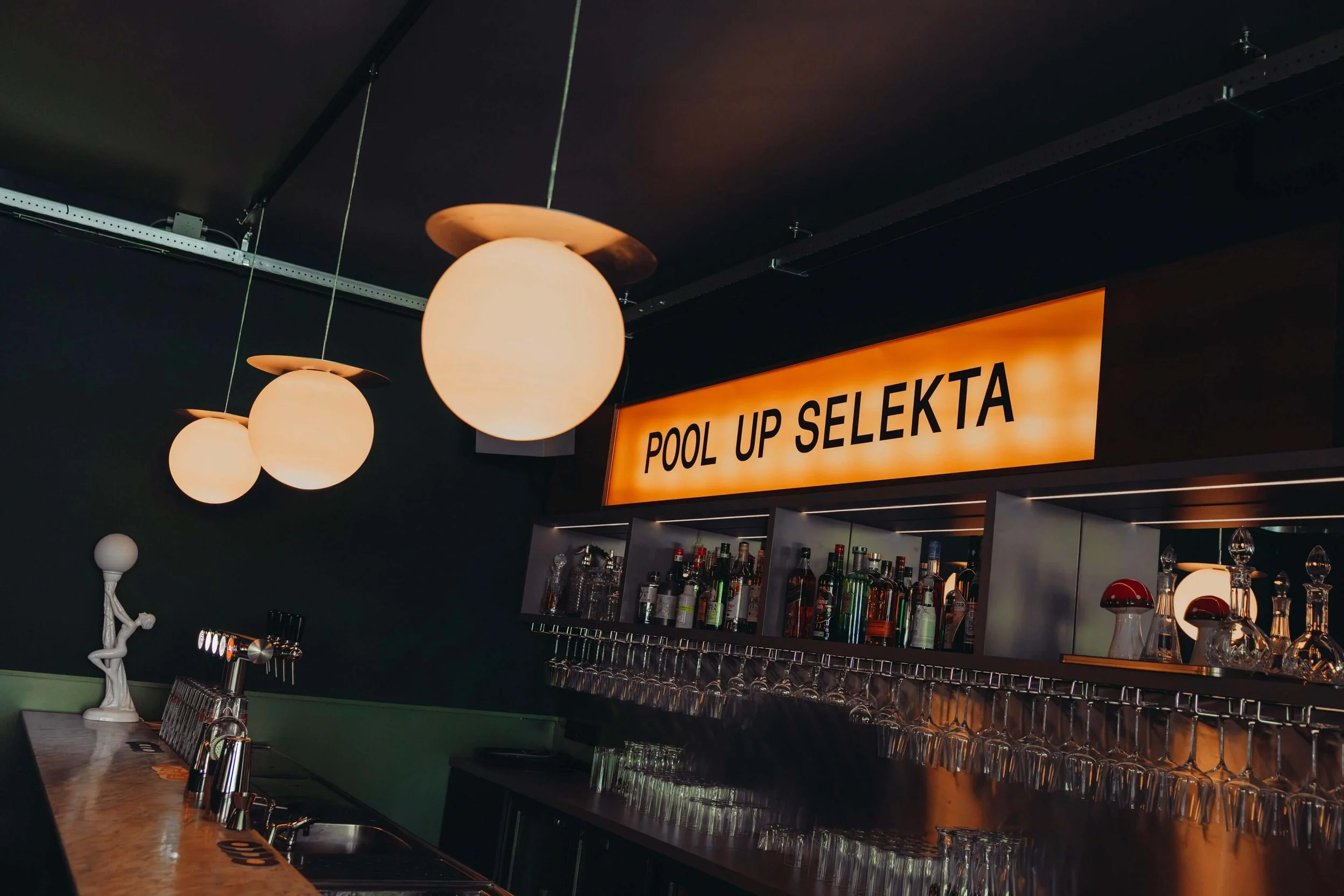 A bar area with hanging spherical lamps, a sign reading 'Pool Up Selekta', shelves with liquor bottles, and glasses on the counter.