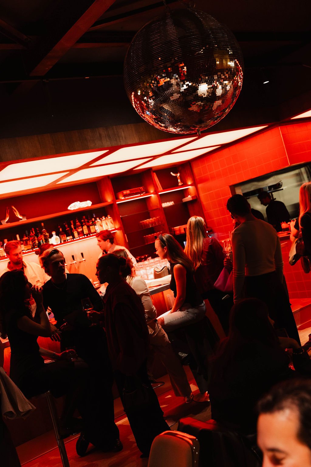 People socializing at a bar with red lighting and a disco ball hanging from the ceiling.