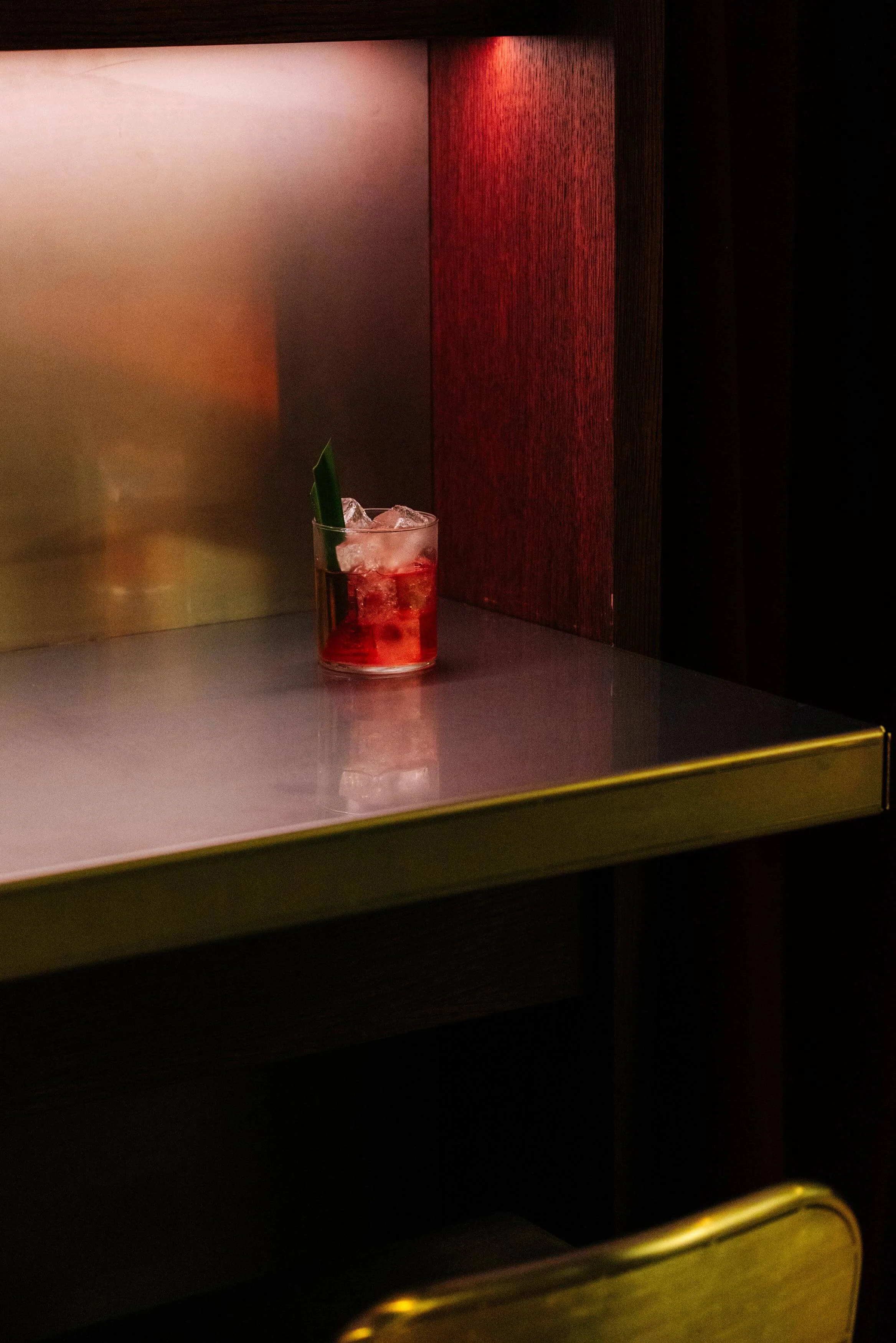 A glass of red cocktail with ice and a green leaf garnish on a white countertop, with a dark red textured background and black curtains.