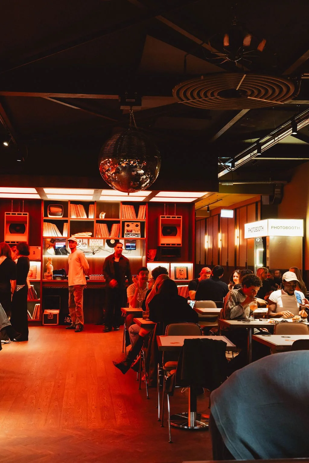A lively indoor scene at a bar or lounge with people sitting at tables and standing near the bar. The warm lighting reflects a cozy atmosphere, with a large disco ball hanging from the ceiling and a bookshelf filled with records and sound equipment b