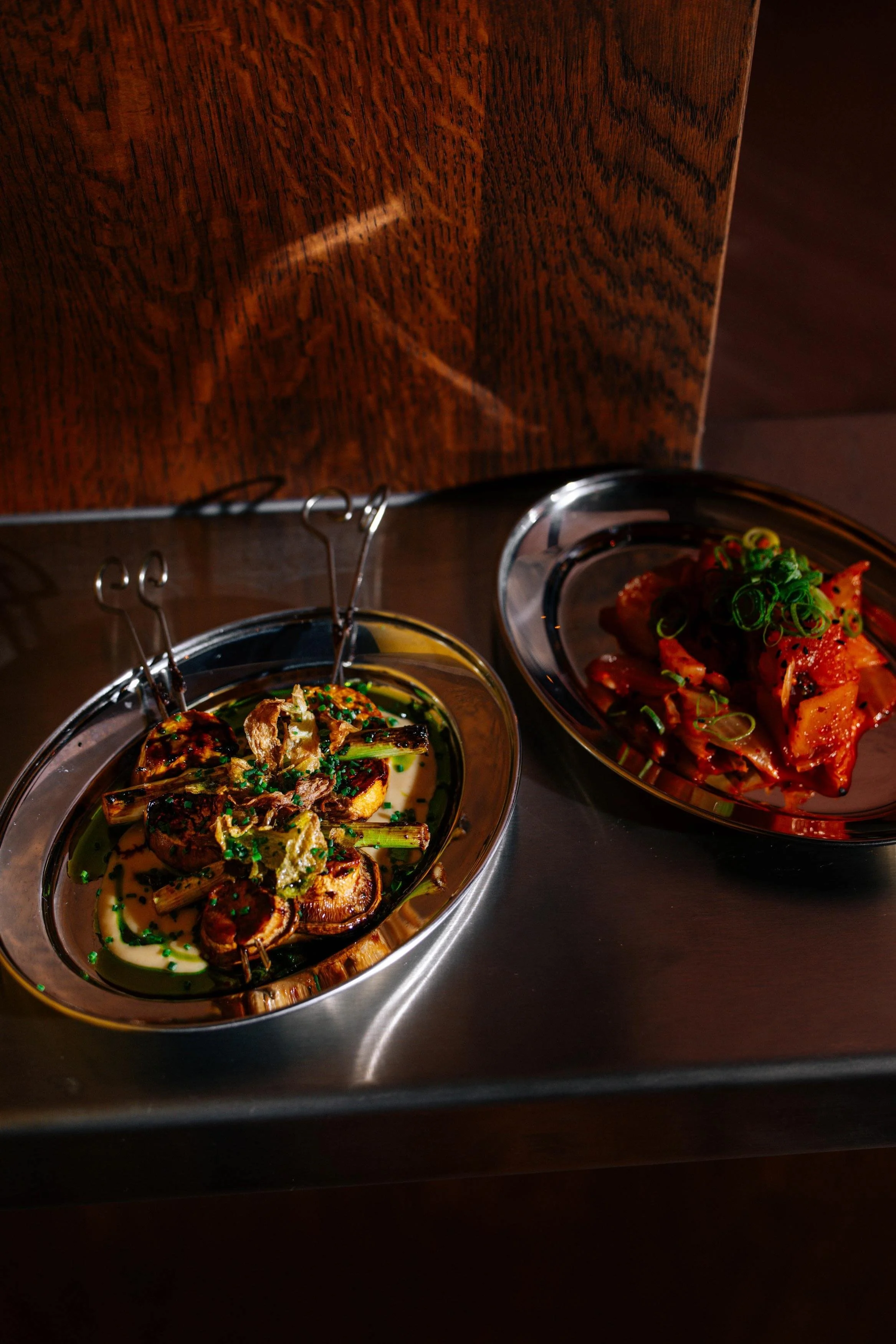 Two dishes of gourmet food on a metal tray, with a dark wood background. One dish contains grilled vegetables with garnish, and the other appears to contain a spicy meat or seafood dish with green onions.