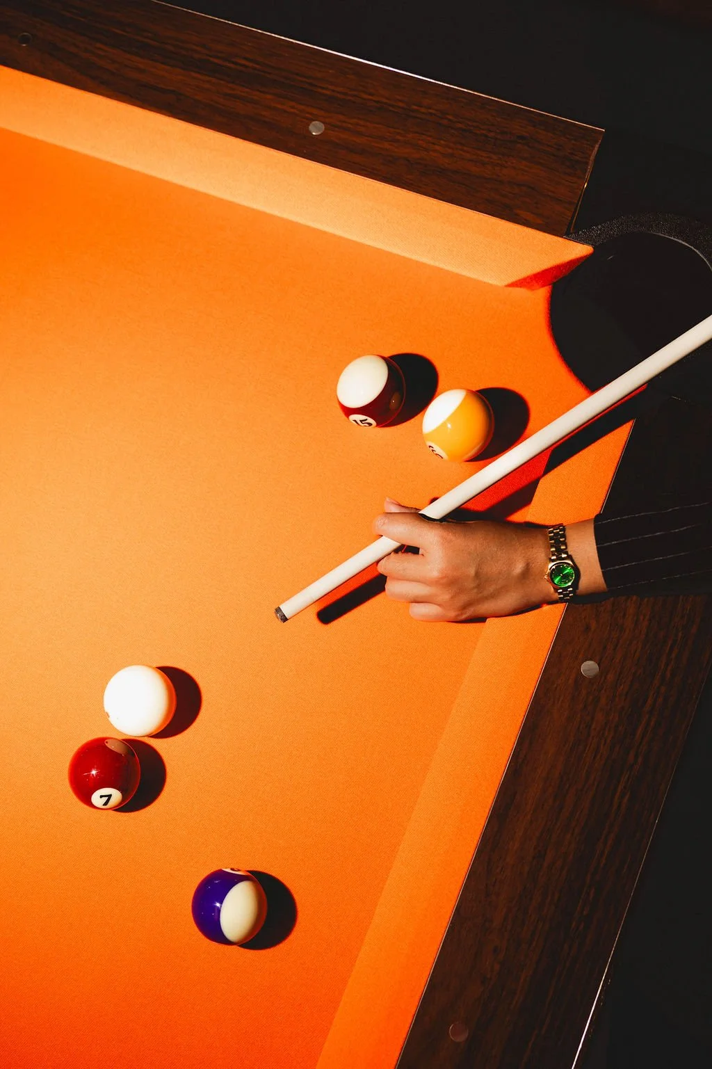 A person with a watch on their left wrist is holding a cue stick on a pool table with orange felt. Four billiard balls are visible on the table: a striped red and white ball numbered 7, a solid yellow ball, a solid white ball, and a purple and white 