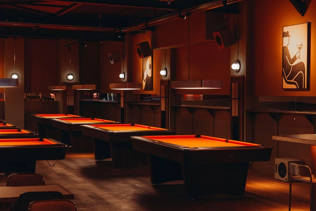 An empty room with multiple pool tables illuminated by warm lighting, decorated with modern artwork and wooden accents.