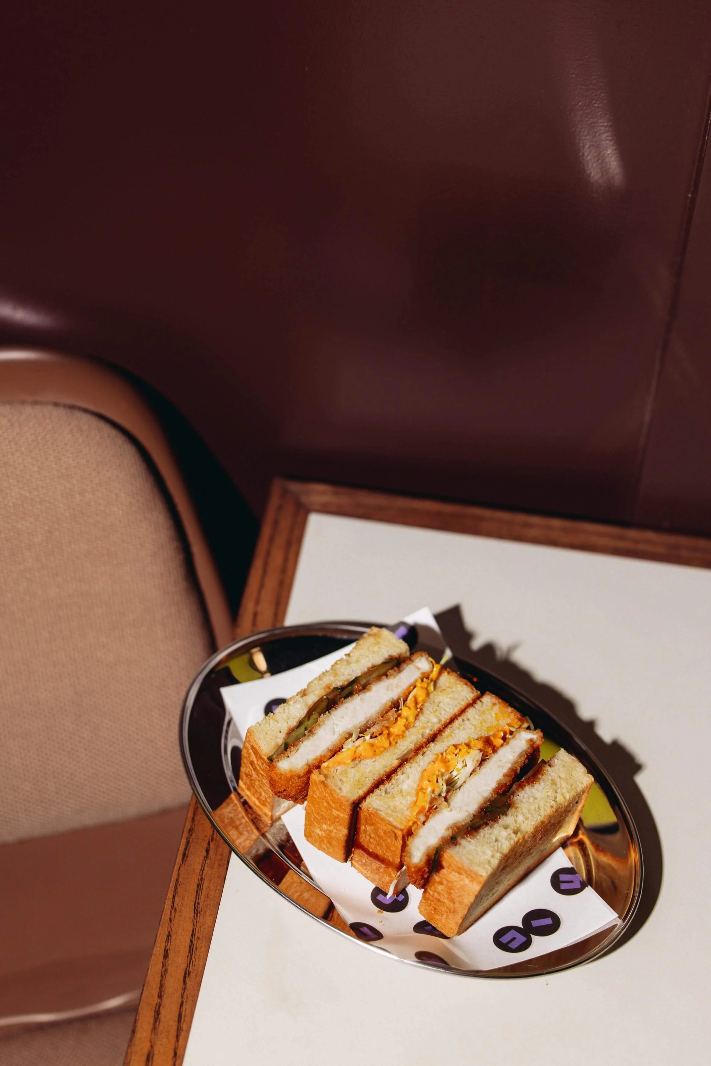 A tray of club sandwiches with layers of bread, lettuce, tomato, cheese, and deli meats, served on a black tray lined with white paper that has black and purple circles.