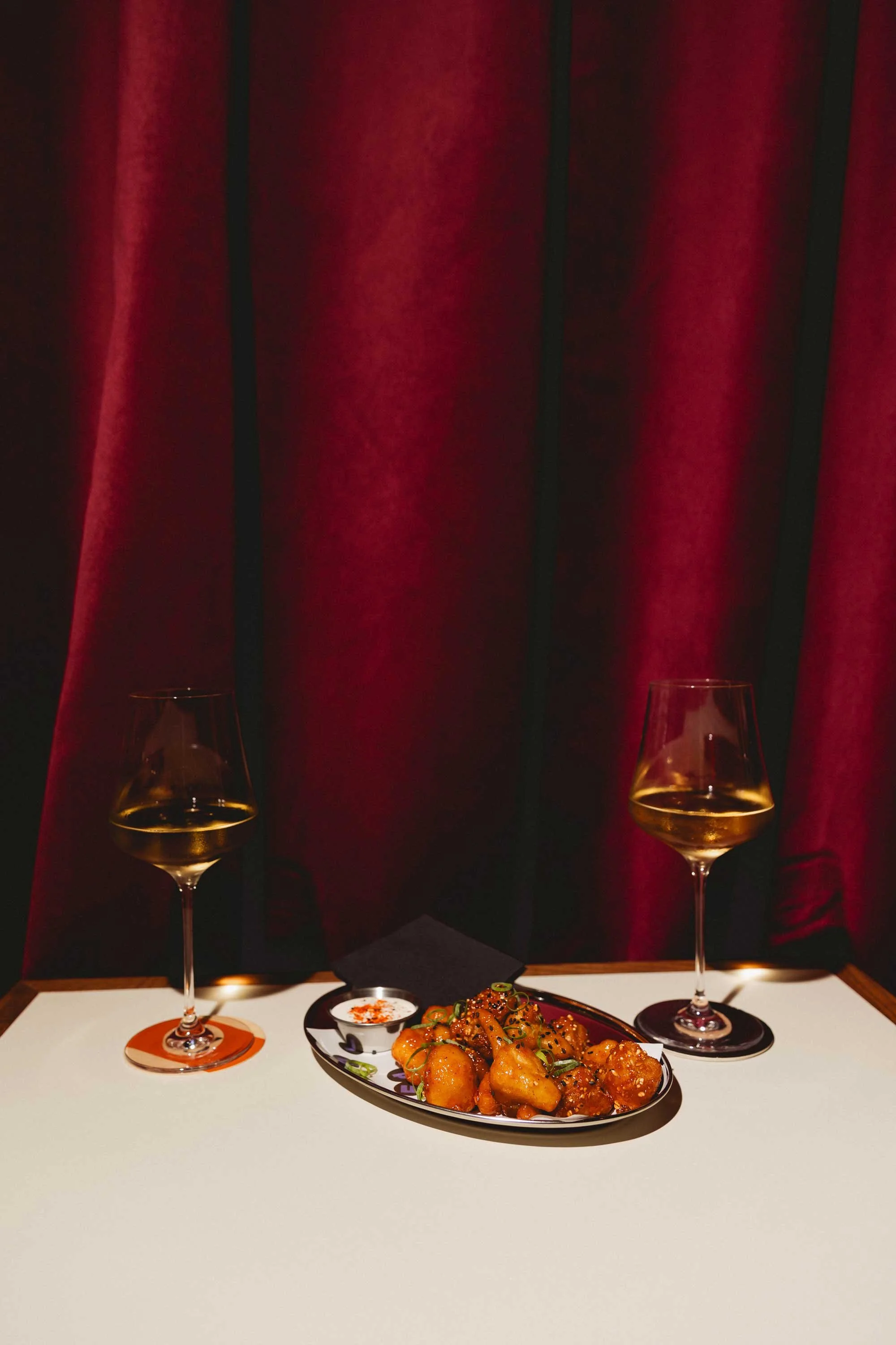 Two glasses of white wine on a table with a plate of Asian-style fried chicken in the foreground, set against a red velvet curtain background.