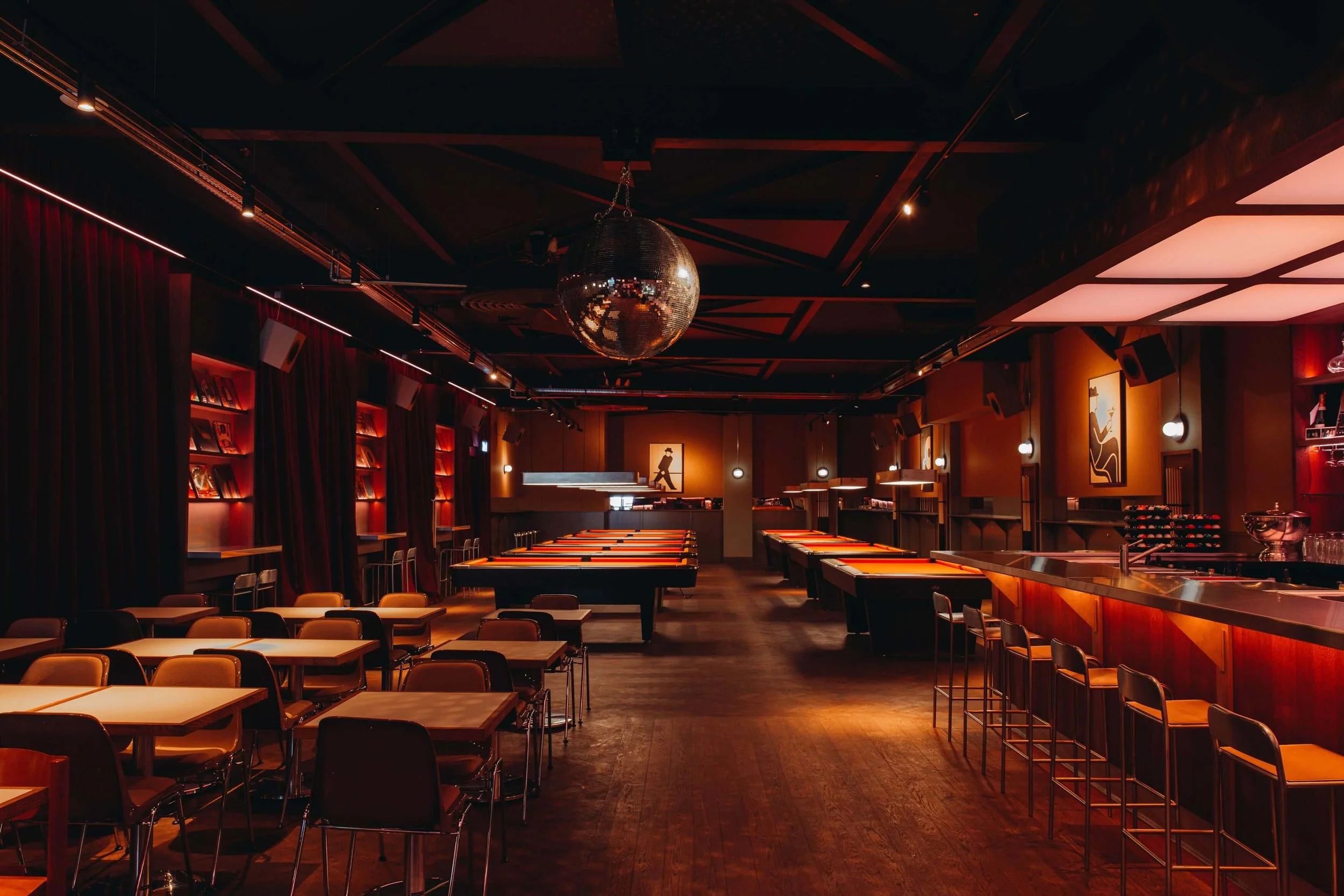 An empty bar or lounge with tables, chairs, a bar counter, and a large disco ball hanging from the ceiling, illuminated with warm, dim lighting.