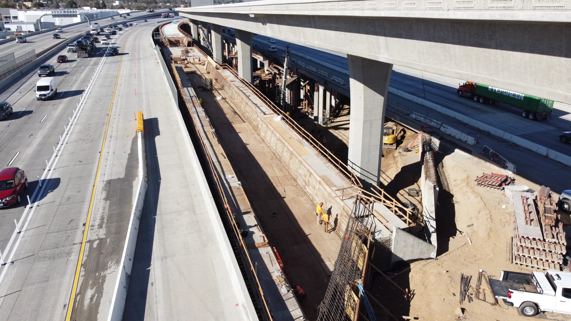 Aerial view of highway construction site, showing multiple lanes of traffic on the left, an under-construction elevated pathway or bridge on the right, and construction workers and equipment working on the site.